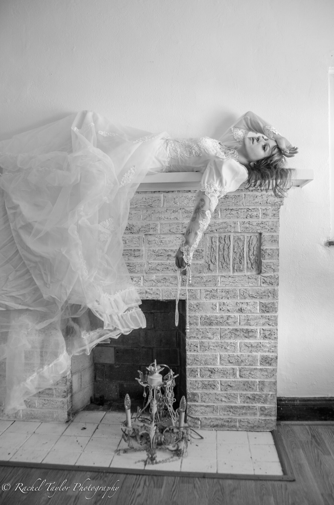 Woman in a flowing dress lying on a brick fireplace mantel, with flowers on the floor below, in a room with plain wall.