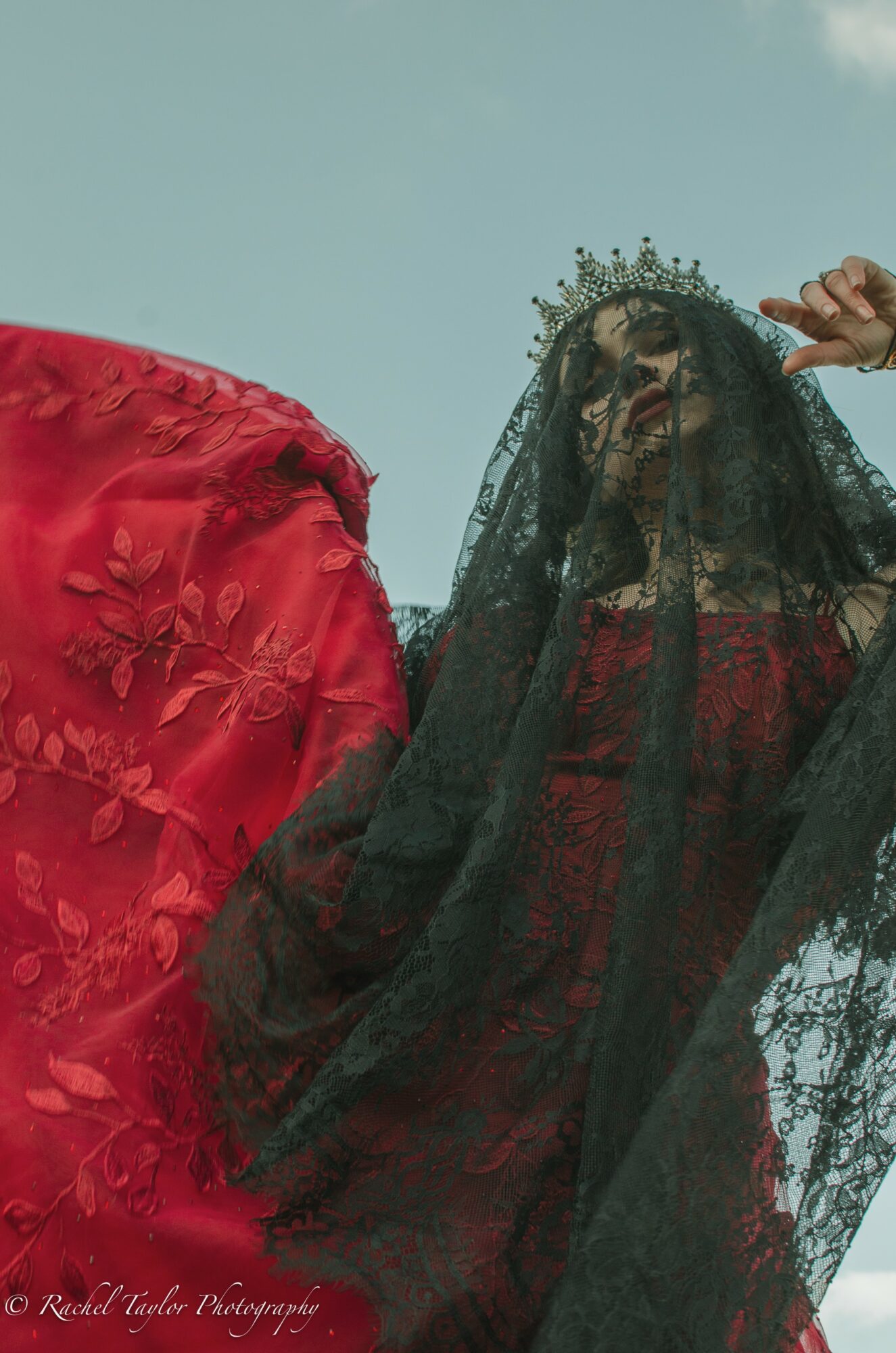 Woman wearing a crown and black veil, dressed in red, with a person in red beside her, against a blue sky.