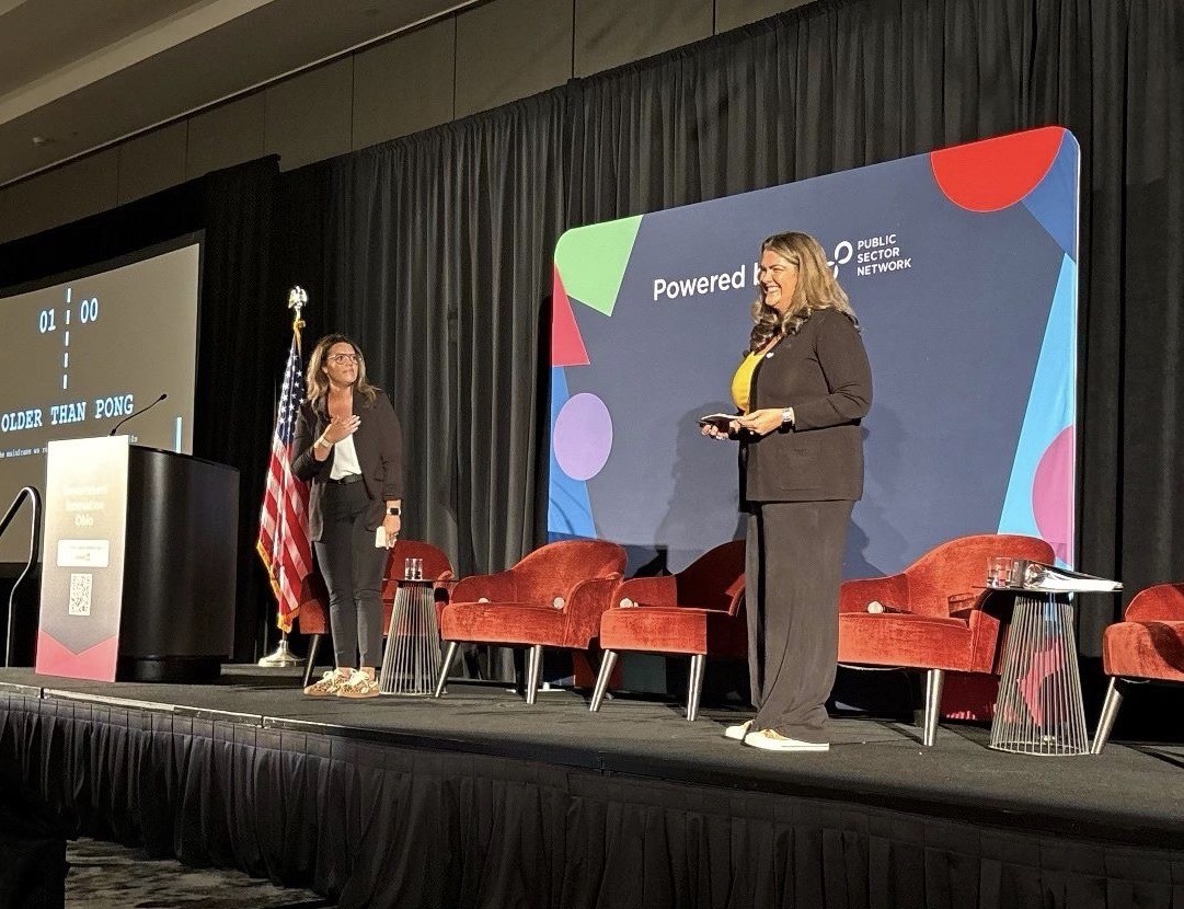 Two women on stage, one speaking at a podium, the other holding a tablet, with a large colorful screen behind them.
