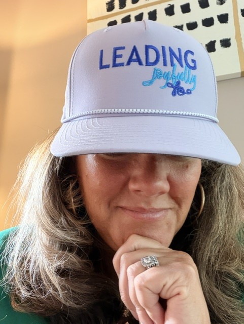 Woman wearing a white cap with blue text and design, resting her chin on her hand, smiling slightly.