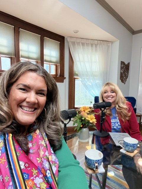Two women smiling at a table with flowers, cups, and microphones in a bright room with large windows and curtains.