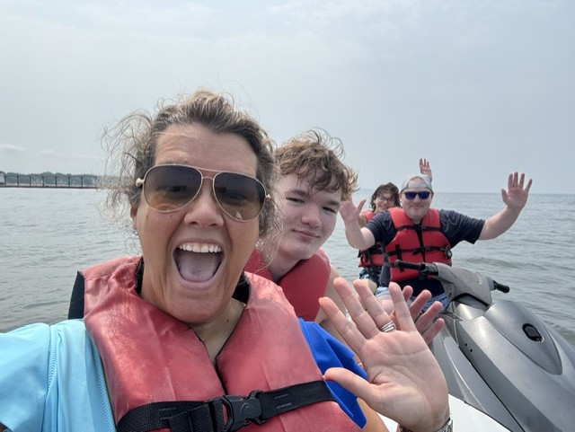 Three people on a boat, smiling and waving, with water and sky in the background.