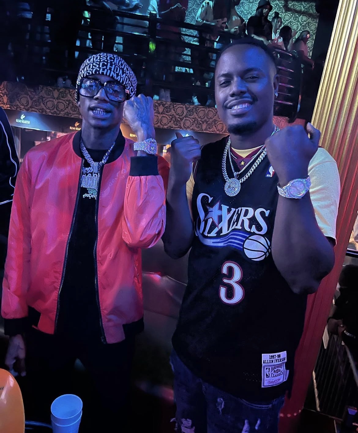 Two men smiling and posing with fists raised in a lively indoor setting with colorful lighting.