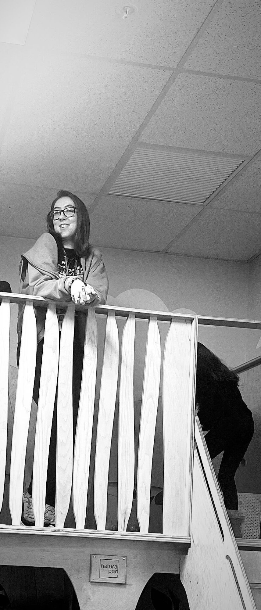 Woman with glasses standing on a balcony railing, smiling, with a person in dark clothing nearby, ceiling with vents above.