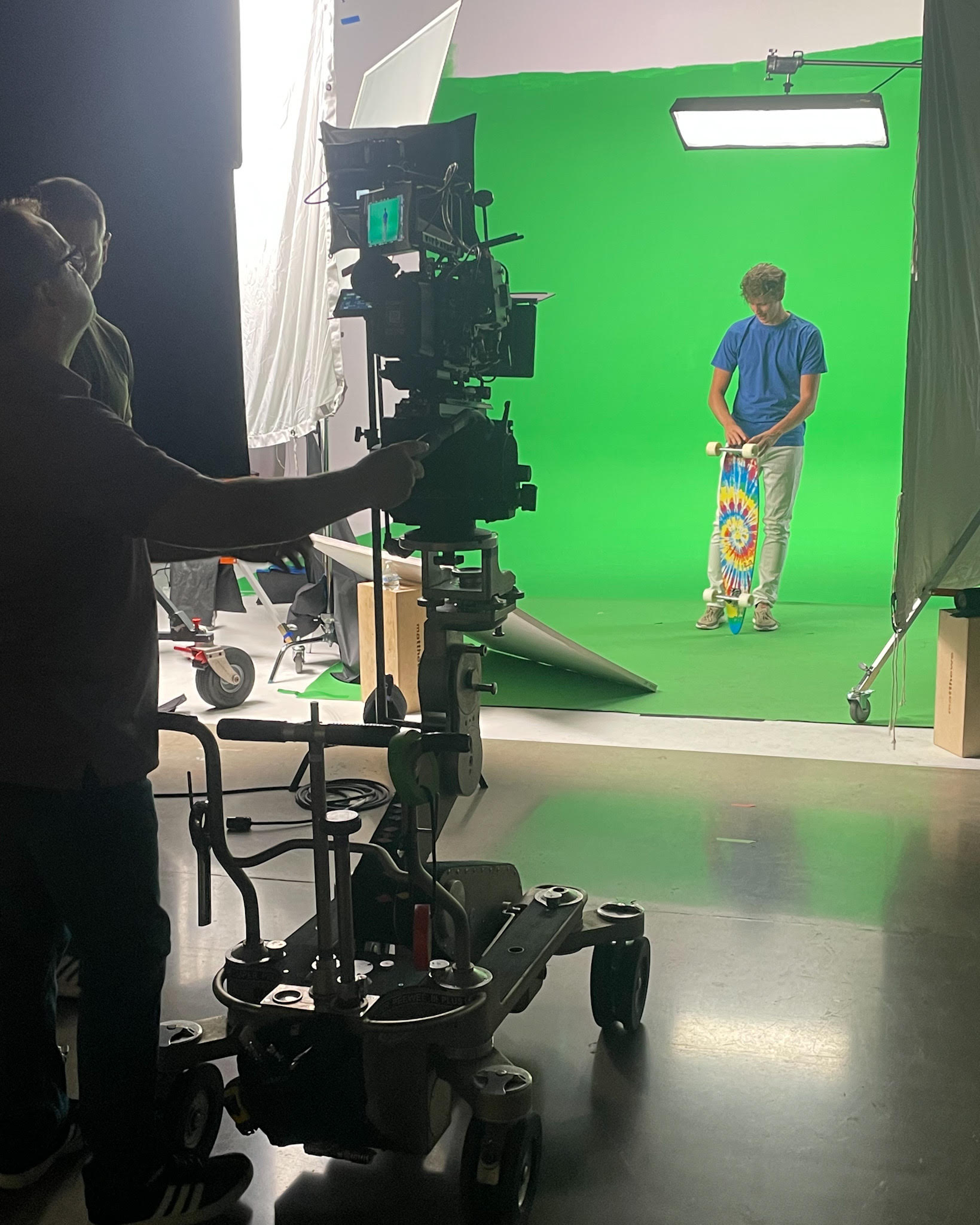 Person operating camera in front of a green screen with a child standing and facing away, holding a colorful skateboard.