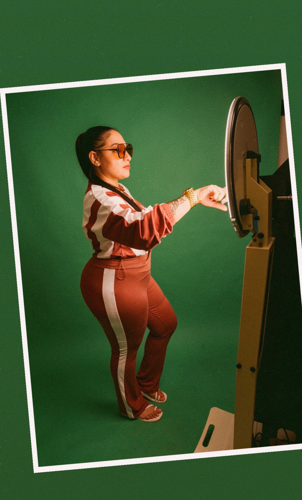 Person in sunglasses and tracksuit using a touchscreen kiosk against a green background.