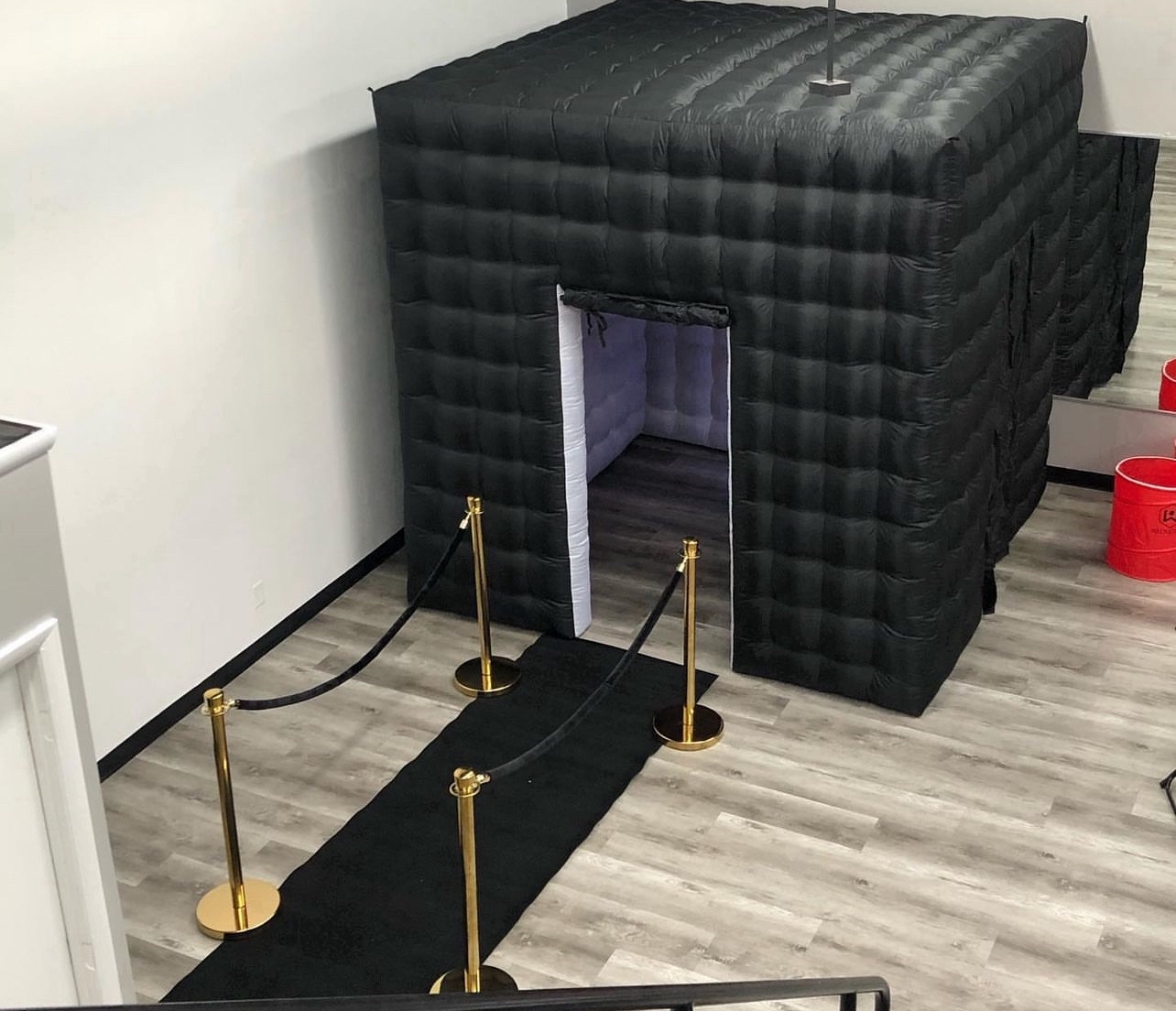 Black inflatable structure with a small entrance, surrounded by a black carpet and gold stanchions, in a room with light-colored flooring.