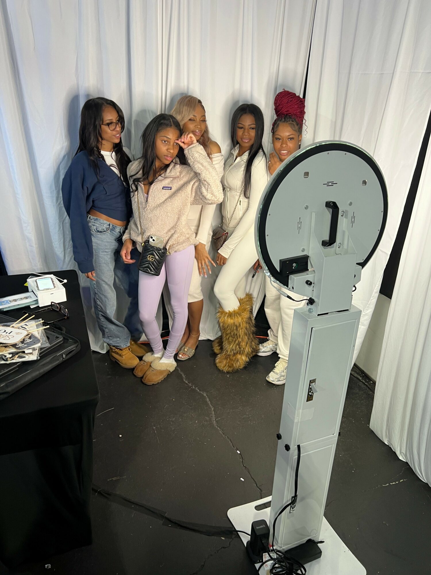 Four women stand together in front of a white curtain, posing for a photo with a large circular device nearby.