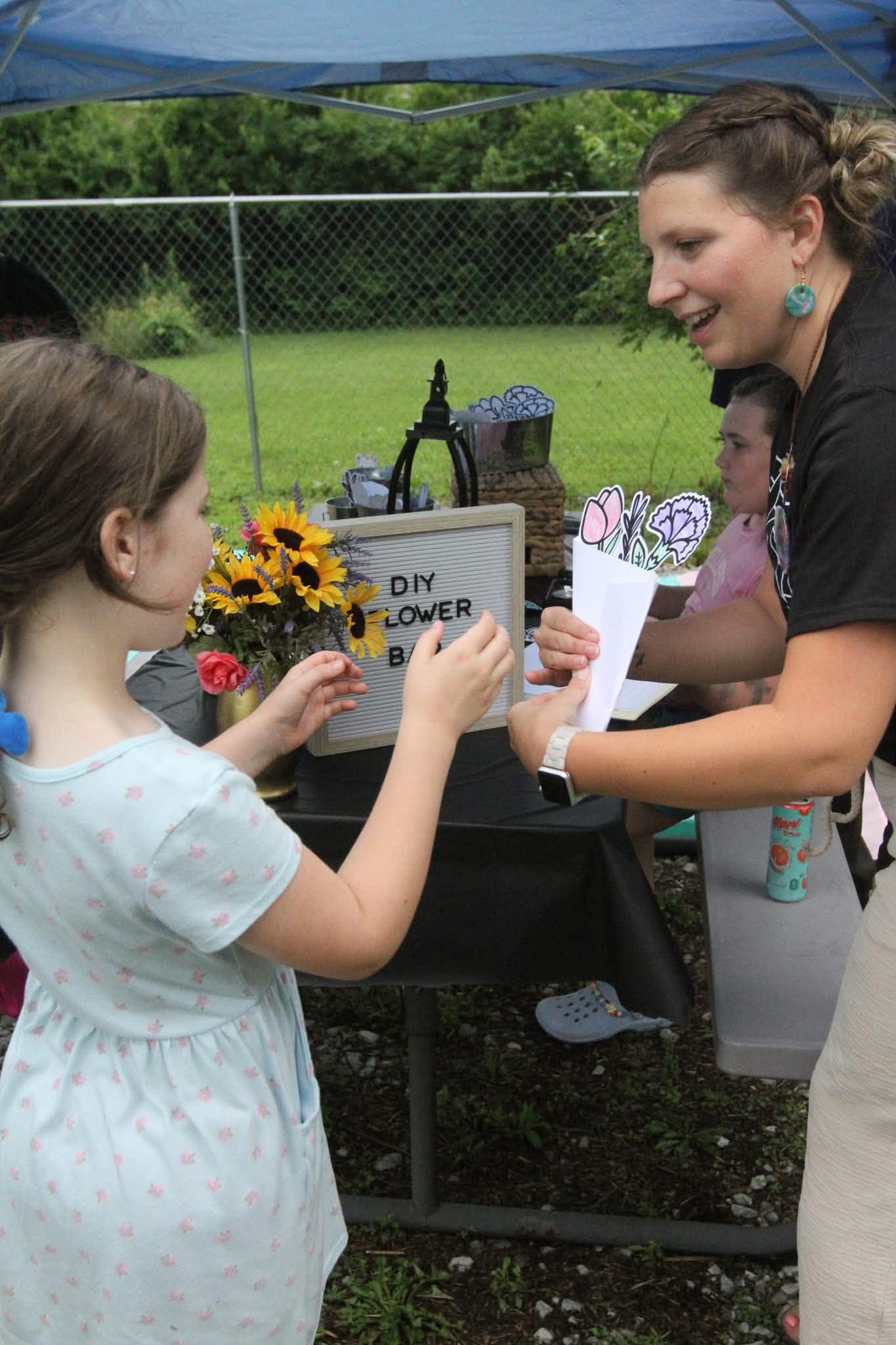 Woman handing a paper to a girl at an outdoor DIY flower stand, with sunflowers and a sign in the background.