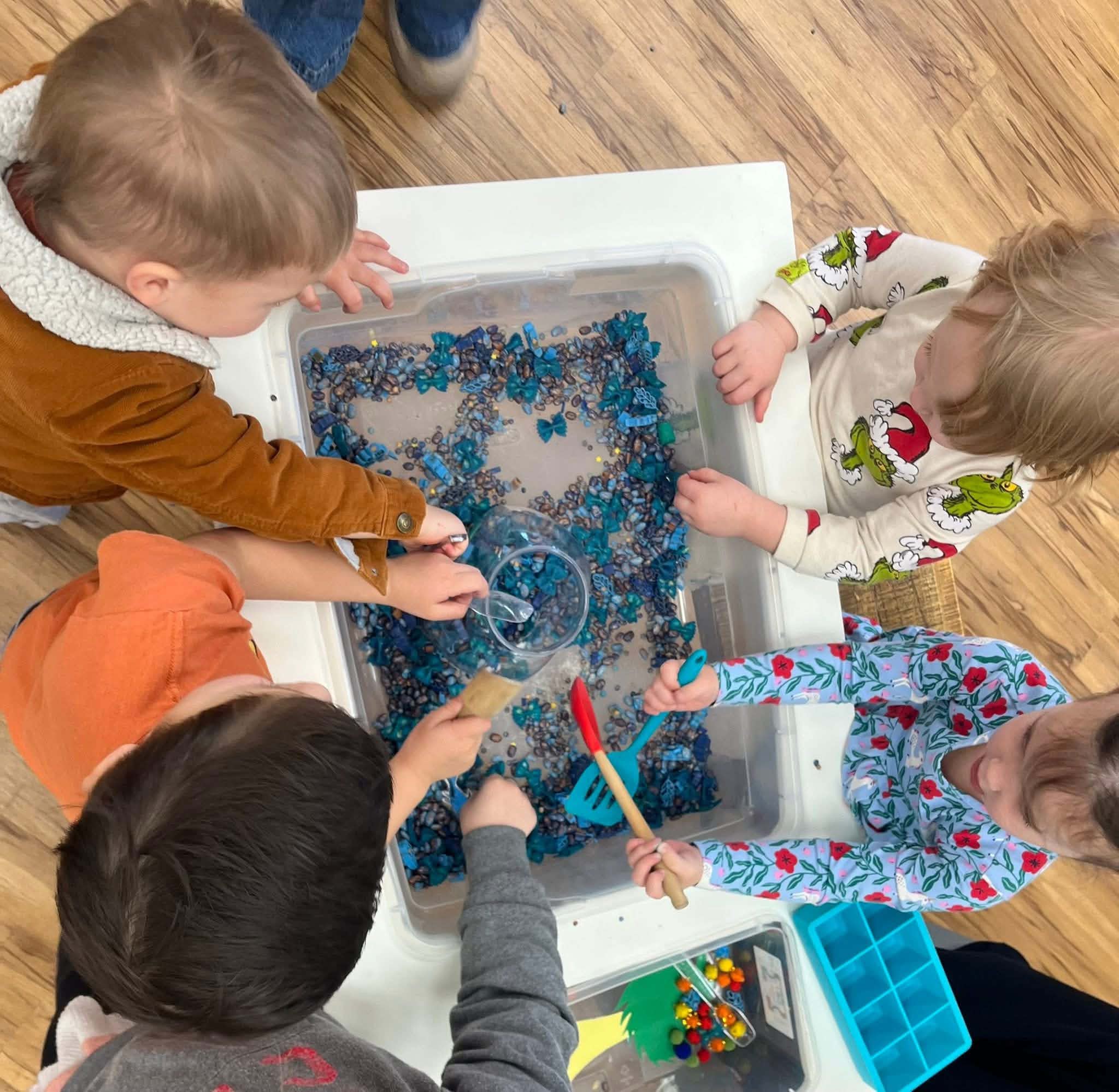 Five children play with blue beads in a clear container on a wooden floor, using tools and containers.