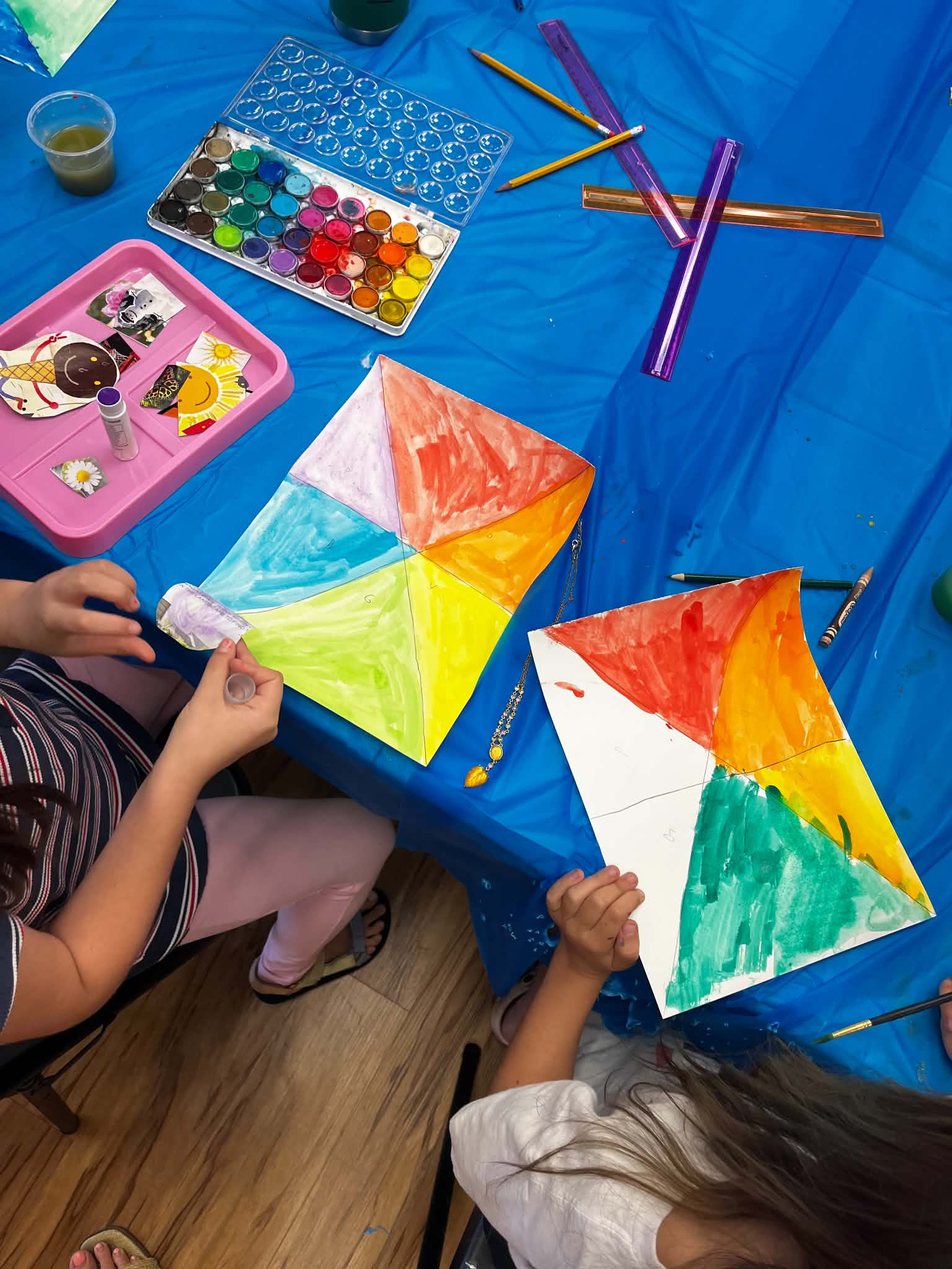 Children painting colorful geometric shapes on paper with paints and brushes, on a blue tablecloth.