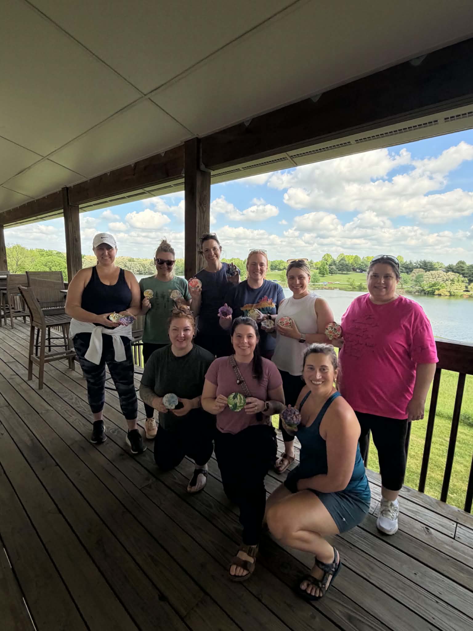 Group of nine people standing on a wooden deck with a scenic view of a lake and blue sky, some holding objects.
