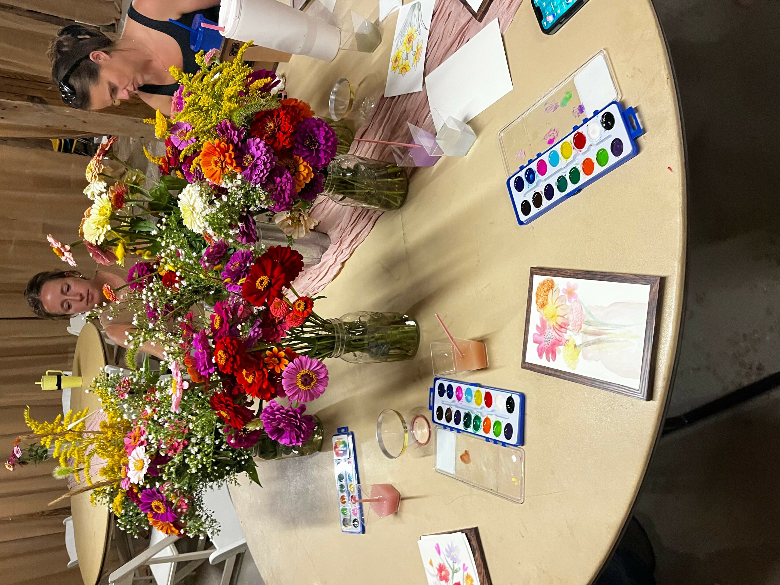 Round table with colorful flower arrangements, watercolor paints, brushes, and paper, with two women in the background.