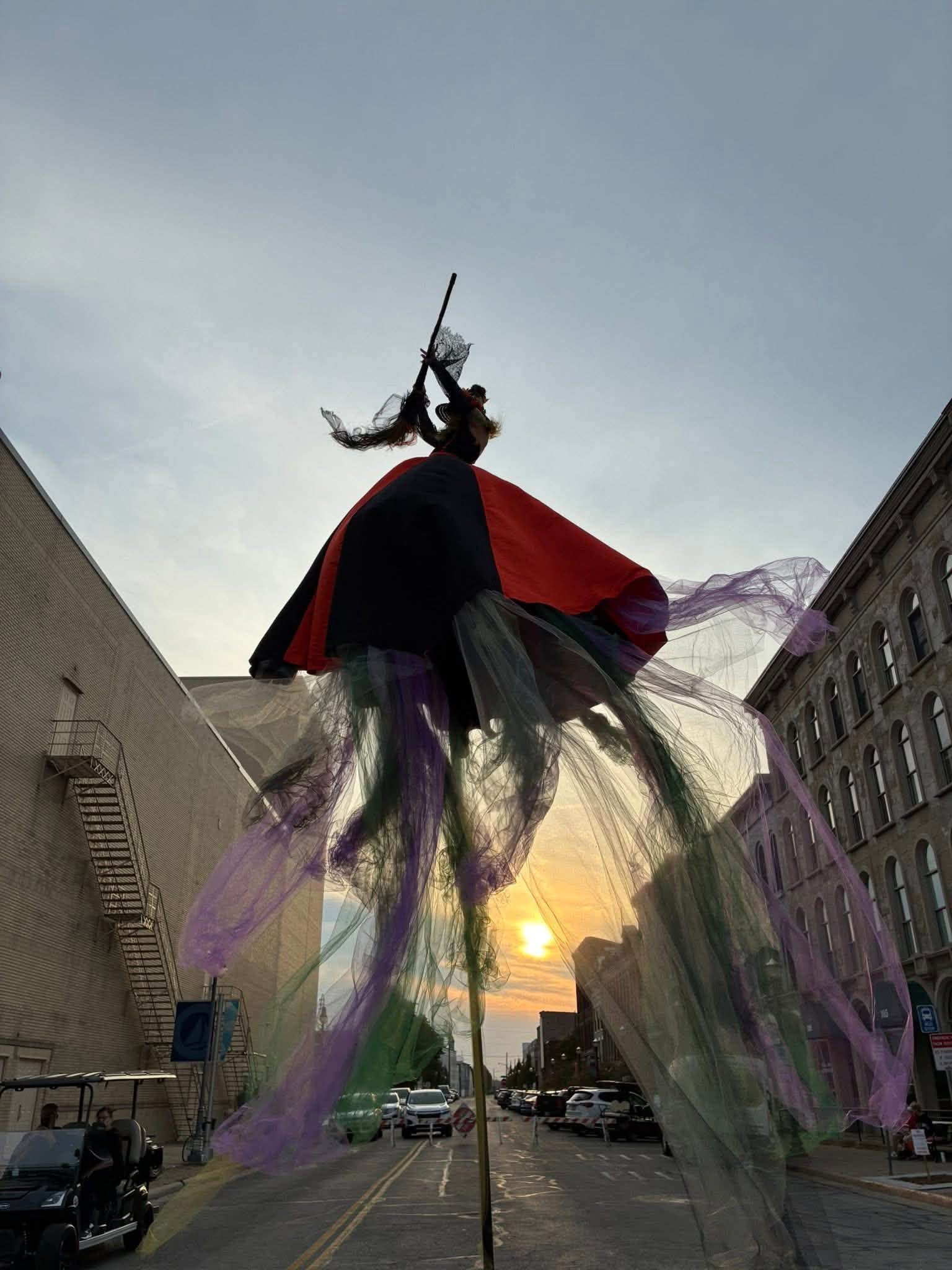 A large puppet or figure with colorful flowing fabric stands on a city street during sunset, with buildings and cars in the background.