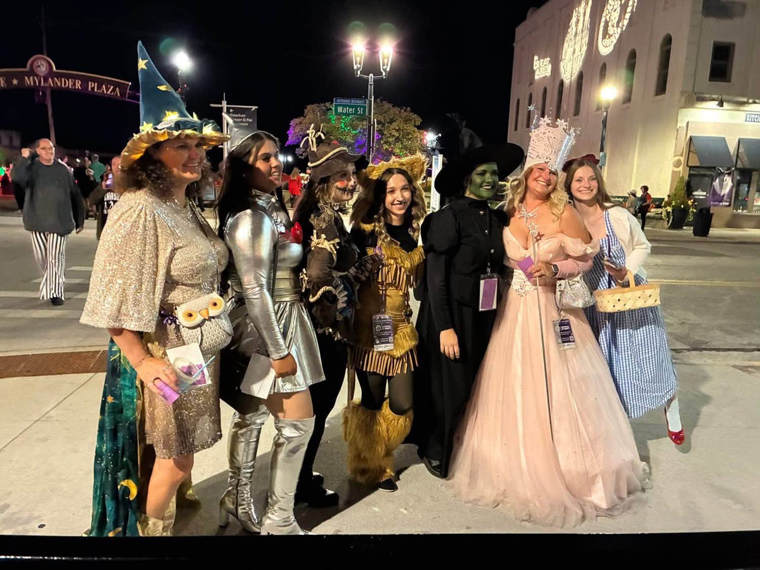 Group of people in costumes standing outdoors at night, some smiling and holding props, with a building and streetlights in background.