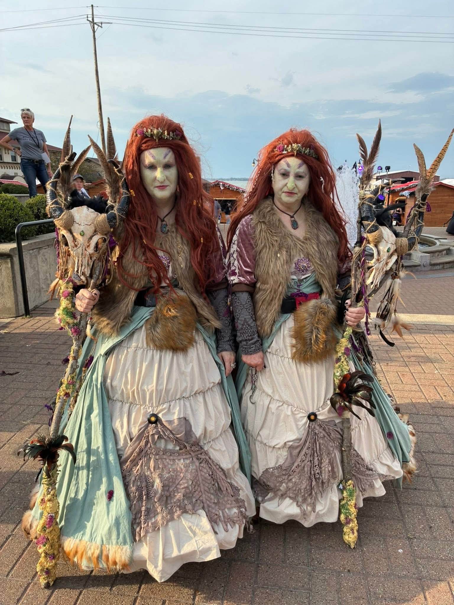 Two people in elaborate costumes with red hair, standing outdoors on a paved area, with a person and vehicles in the background.