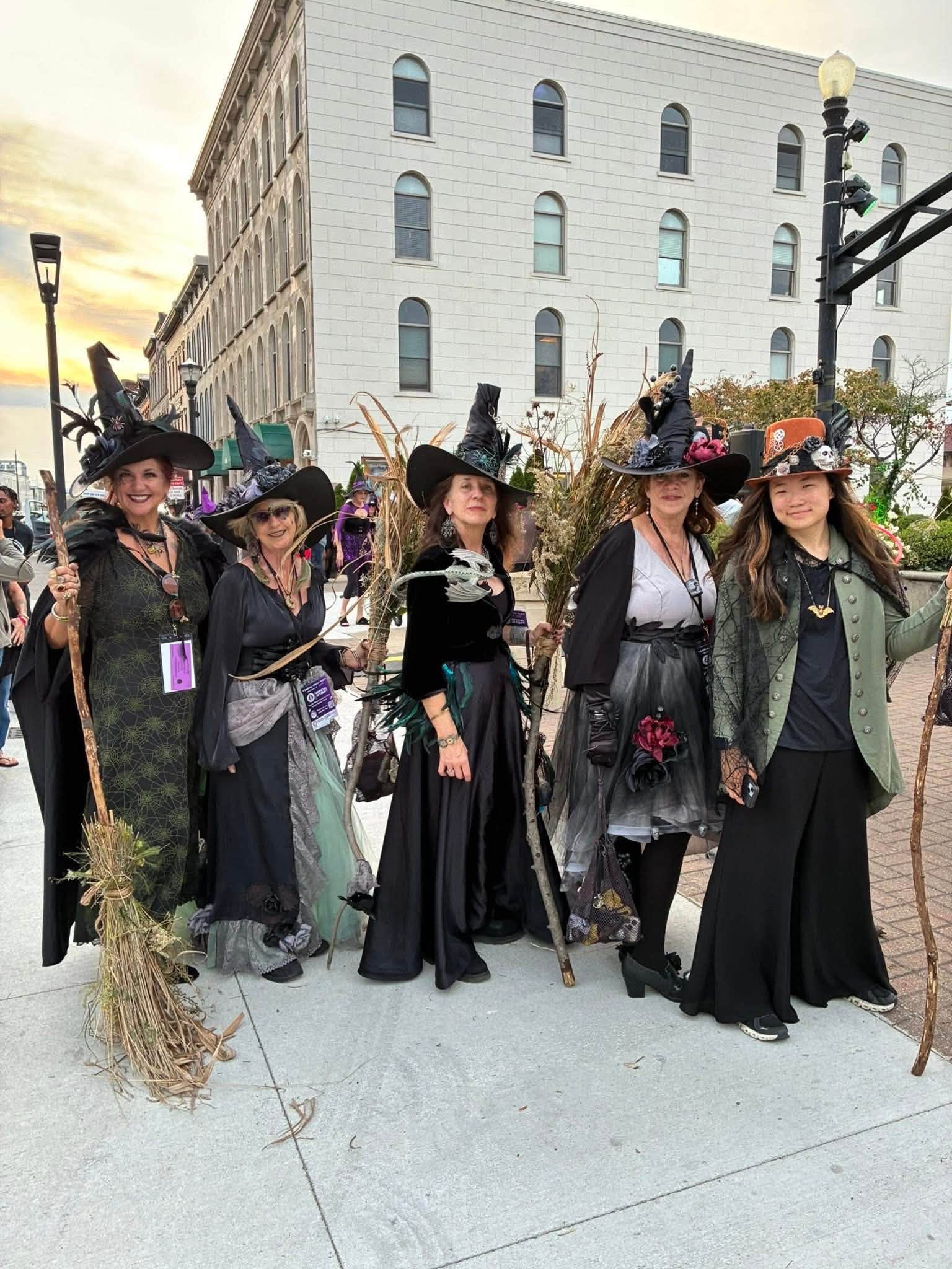 Group of women dressed as witches with hats and costumes, standing on a city sidewalk with buildings and streetlights in background.