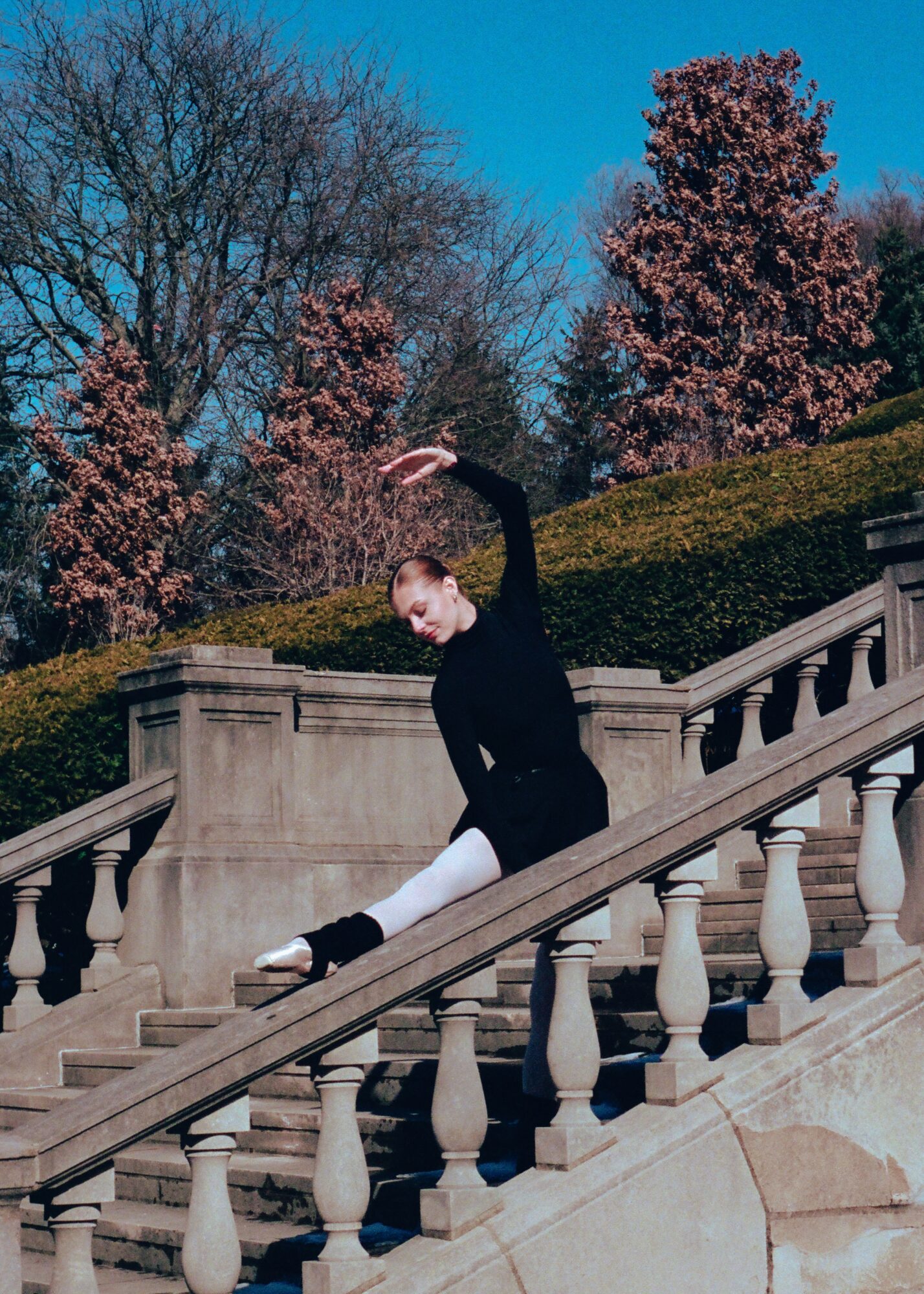 Ballet dancer in black costume stretching on outdoor staircase with trees and pink blossoms in background.
