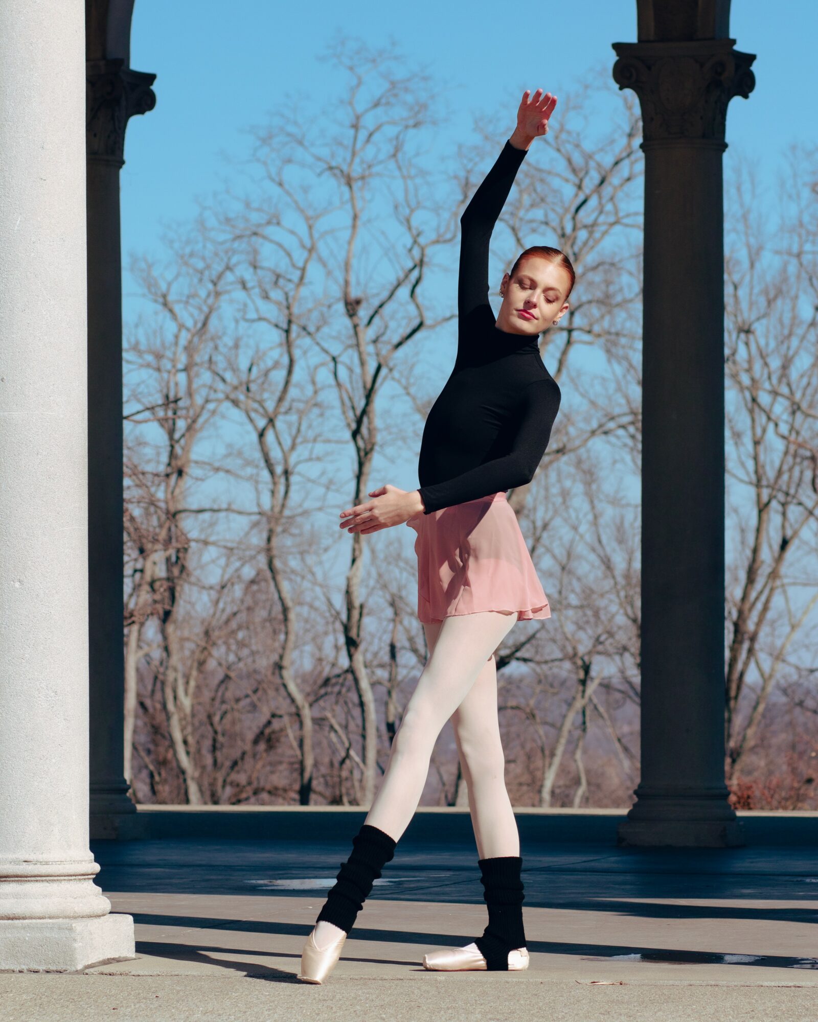 Ballet dancer in black top and pink skirt poses outdoors between columns with trees in background.