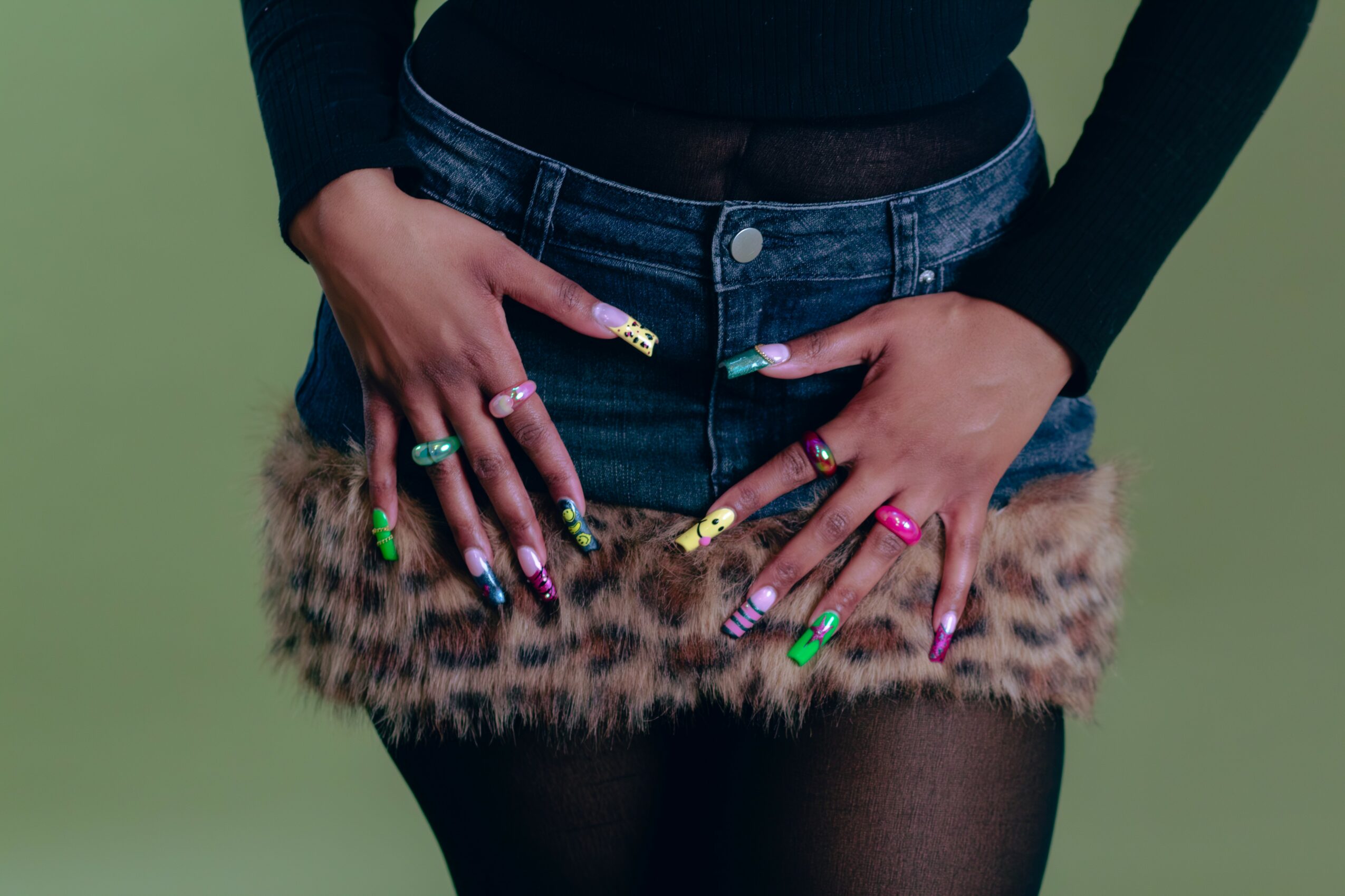 Person wearing a black top, denim shorts with fur trim, and colorful nail polish, standing against a green background.