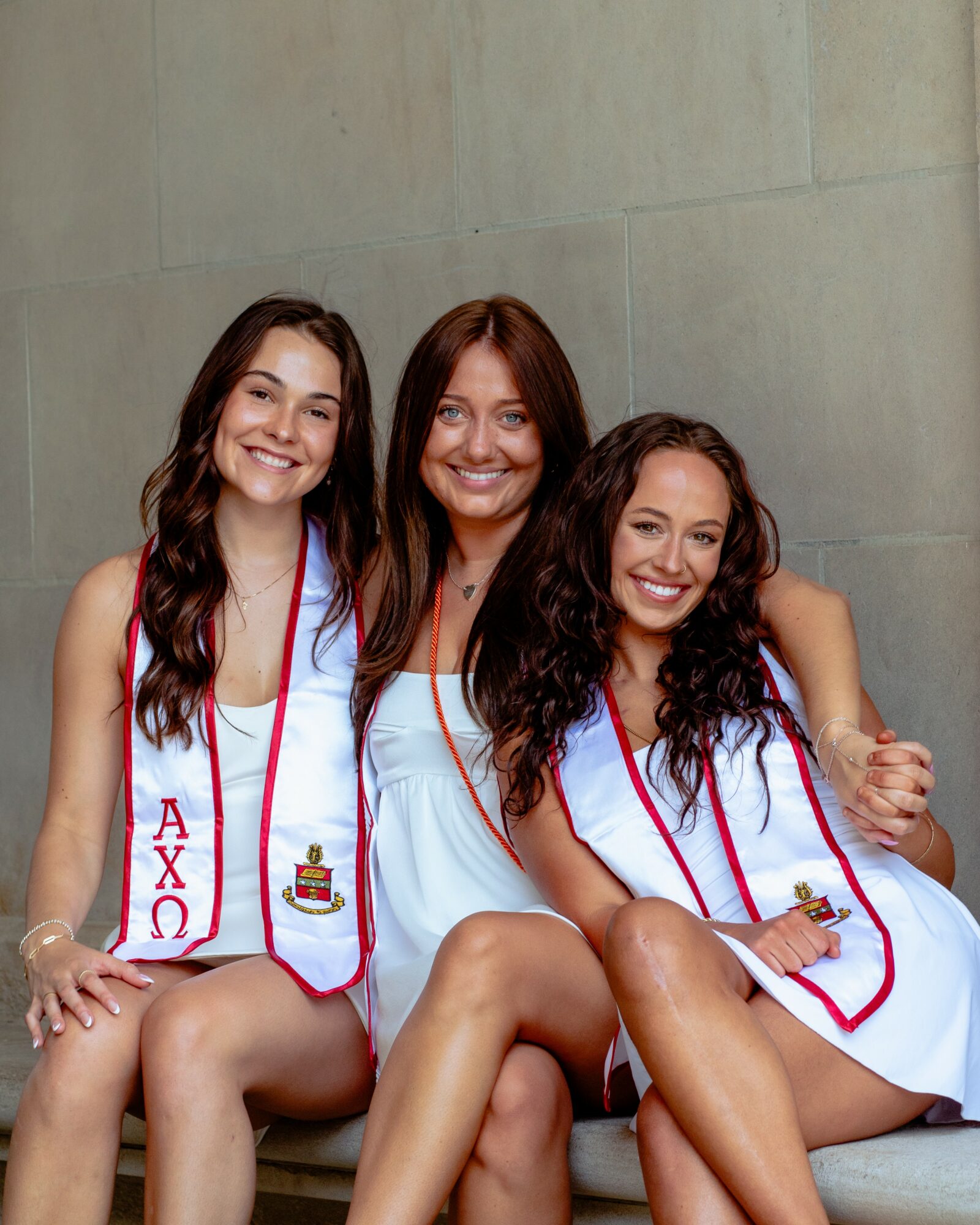 Three women in white graduation gowns sitting together, smiling, with arms around each other, against a plain wall.