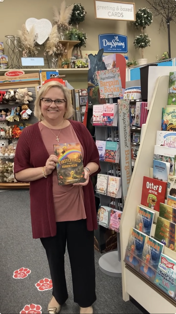 Woman with glasses holding a book in a store with greeting cards and holiday decorations behind her.