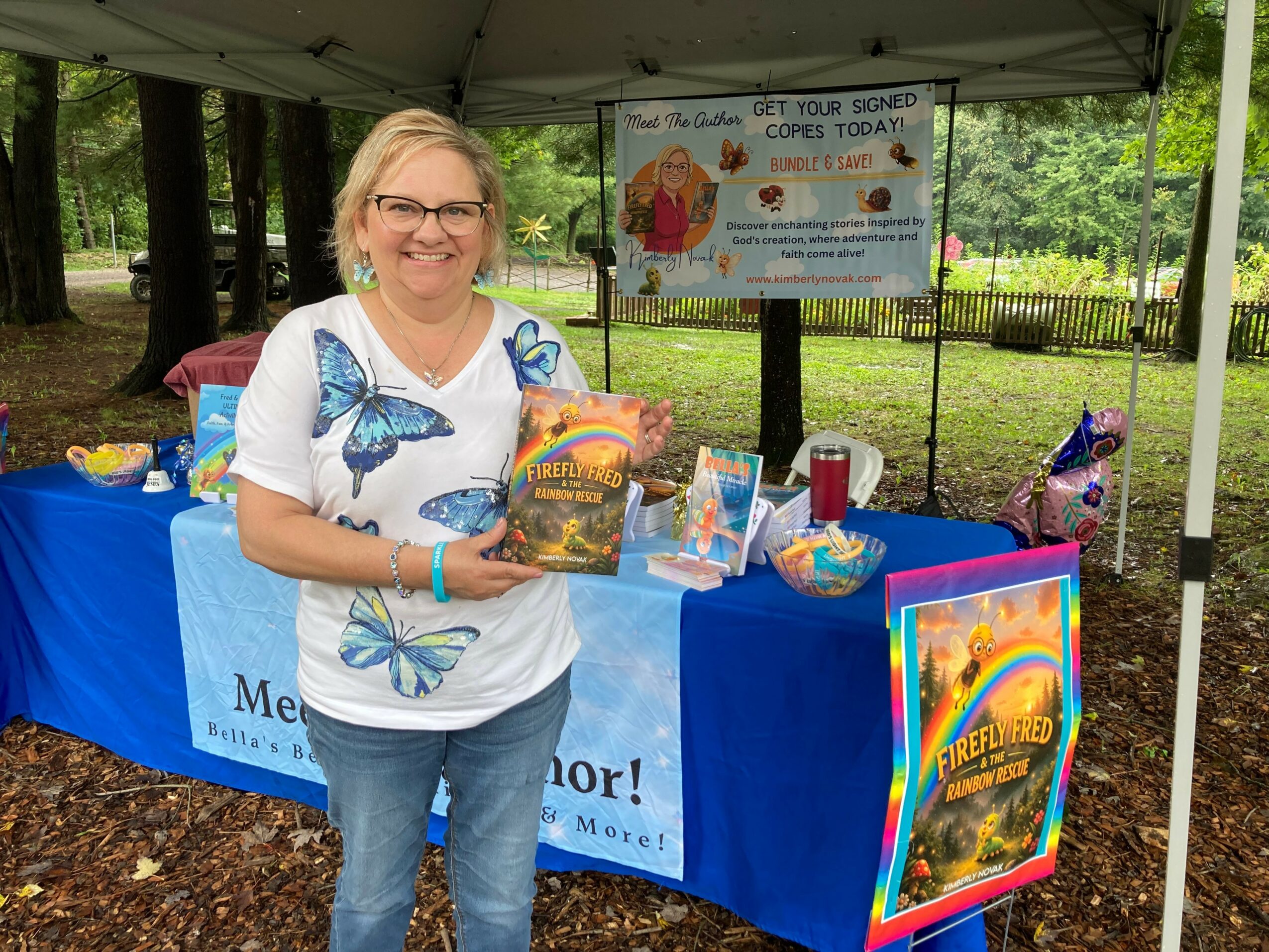 Smiling woman holding a book at outdoor event booth with colorful posters and items, trees and a park in background.