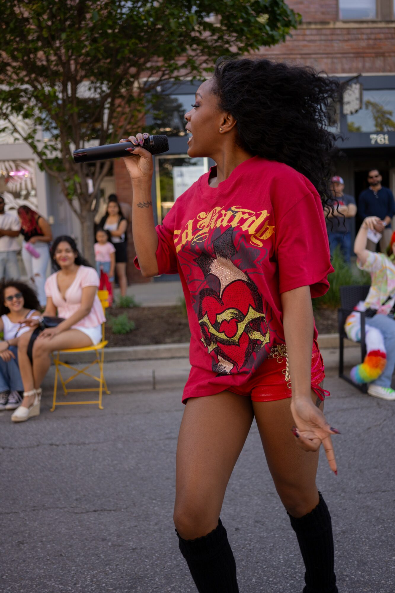 Young woman with curly hair singing into a microphone outdoors, wearing a red T-shirt and shorts, with people sitting and standing nearby.