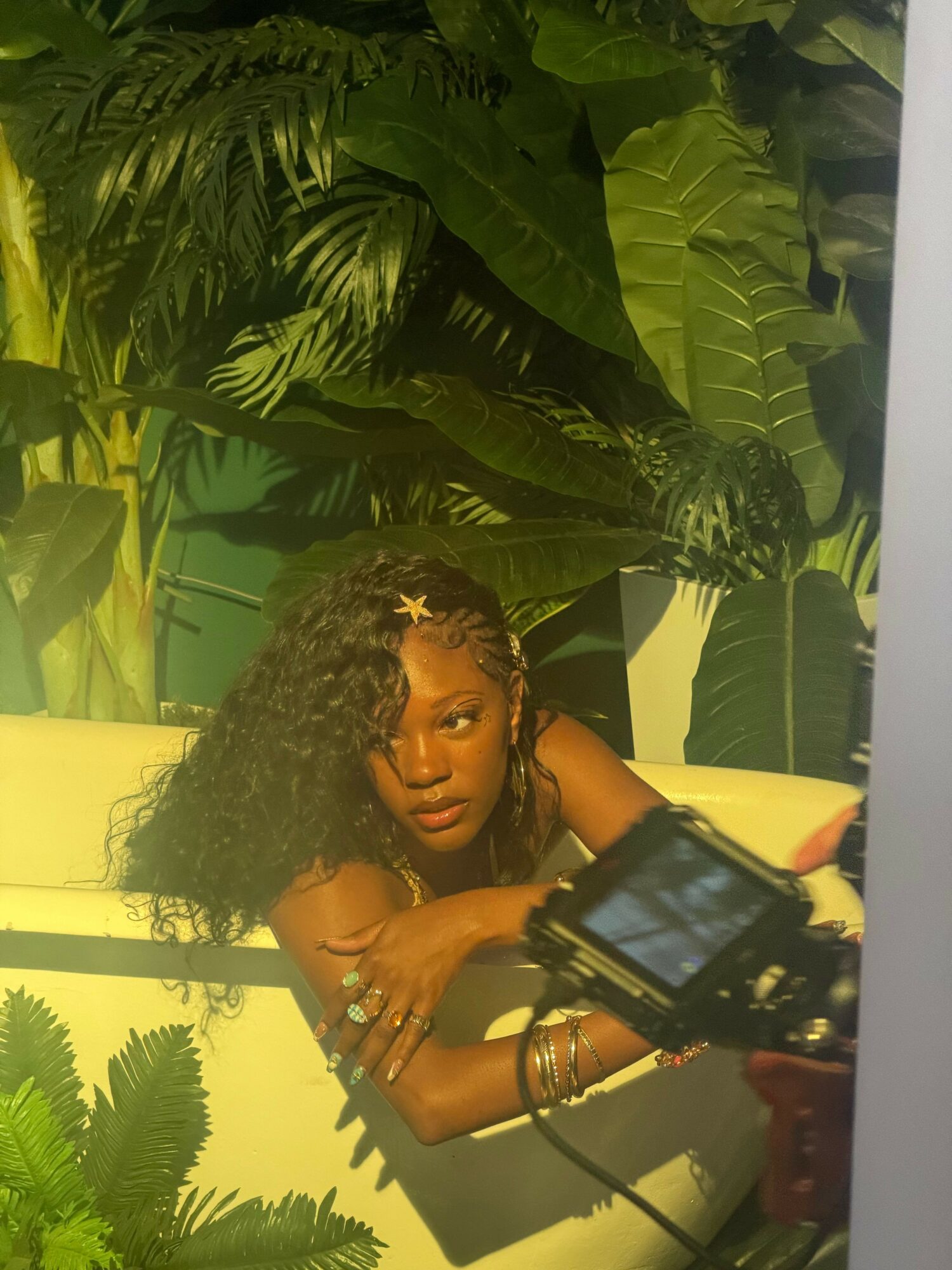 Woman with curly hair and jewelry lying on a yellow surface among large green tropical plants.