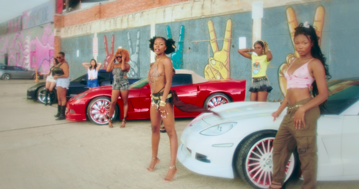Six women stand in front of colorful graffiti and two sports cars, one red and one white, in an urban setting.