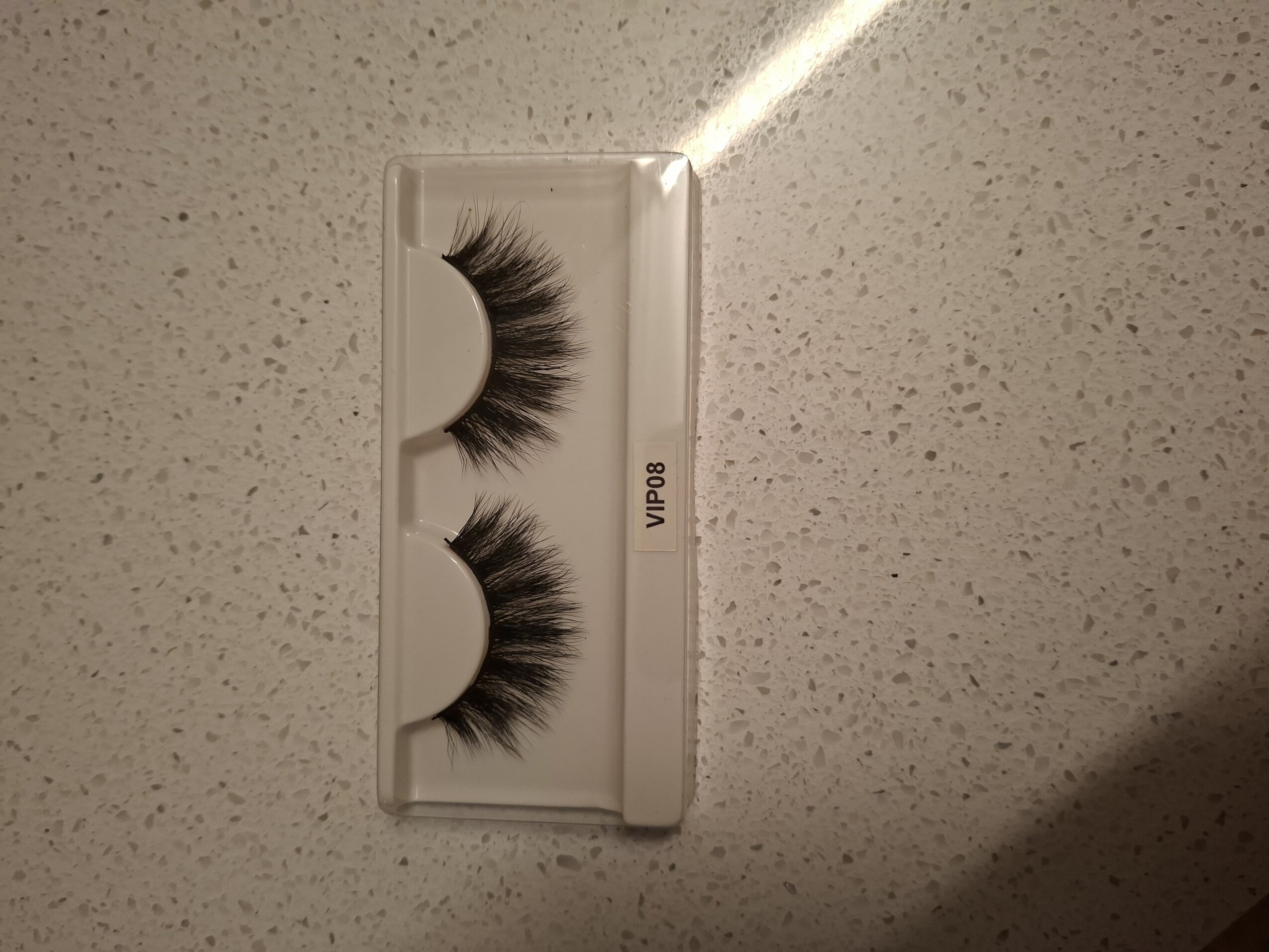 Box of false eyelashes on a speckled surface, with a light reflection above, and a shadow below.