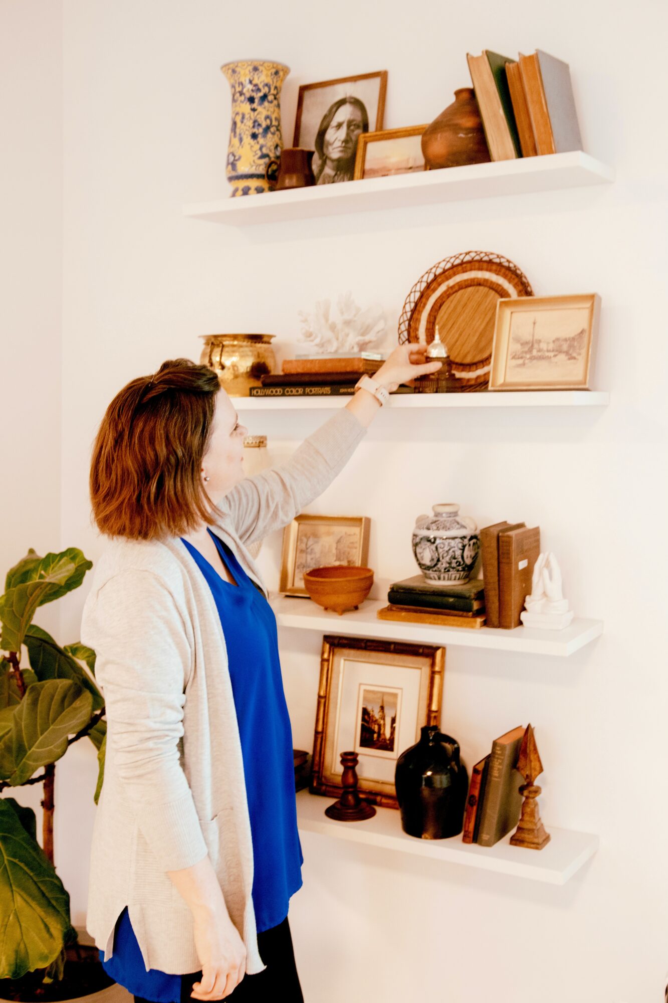 Woman reaching for a decorative object on a white wall shelf with books and framed pictures, in a room with a plant.