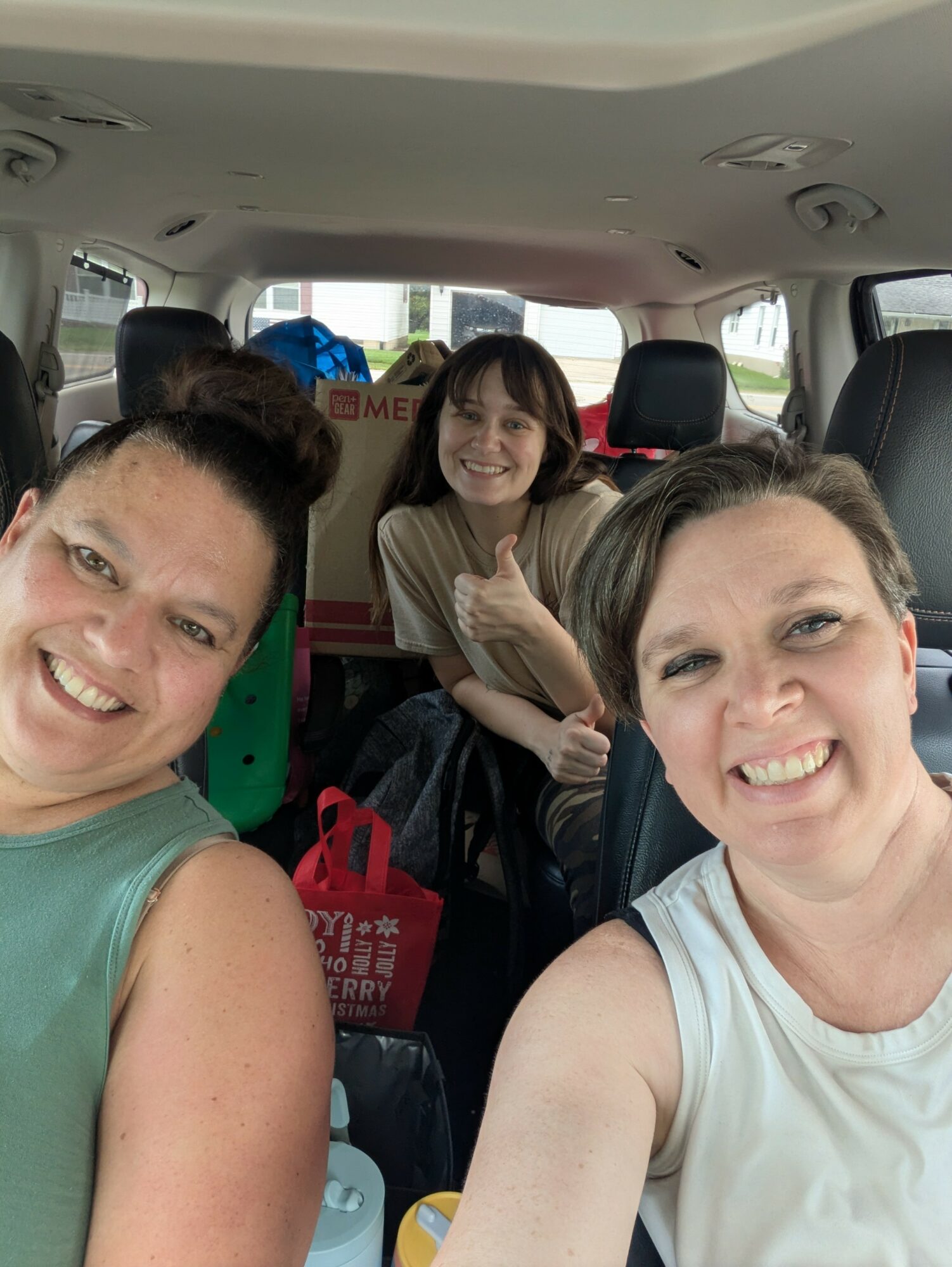 Three women smiling inside a vehicle, with bags and boxes in the background.