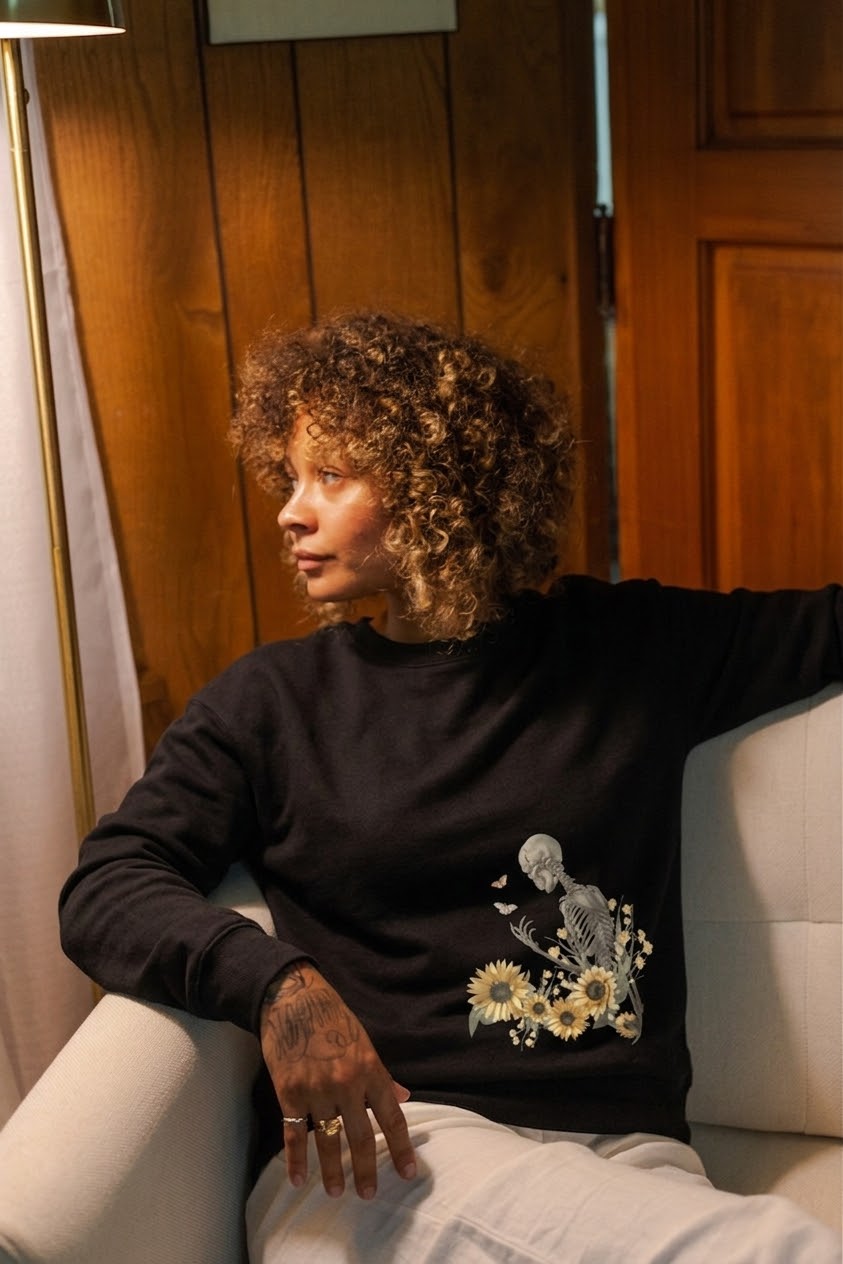 Woman with curly hair sitting on a white couch, wearing a black sweater with floral embroidery, in a wooden-paneled room.