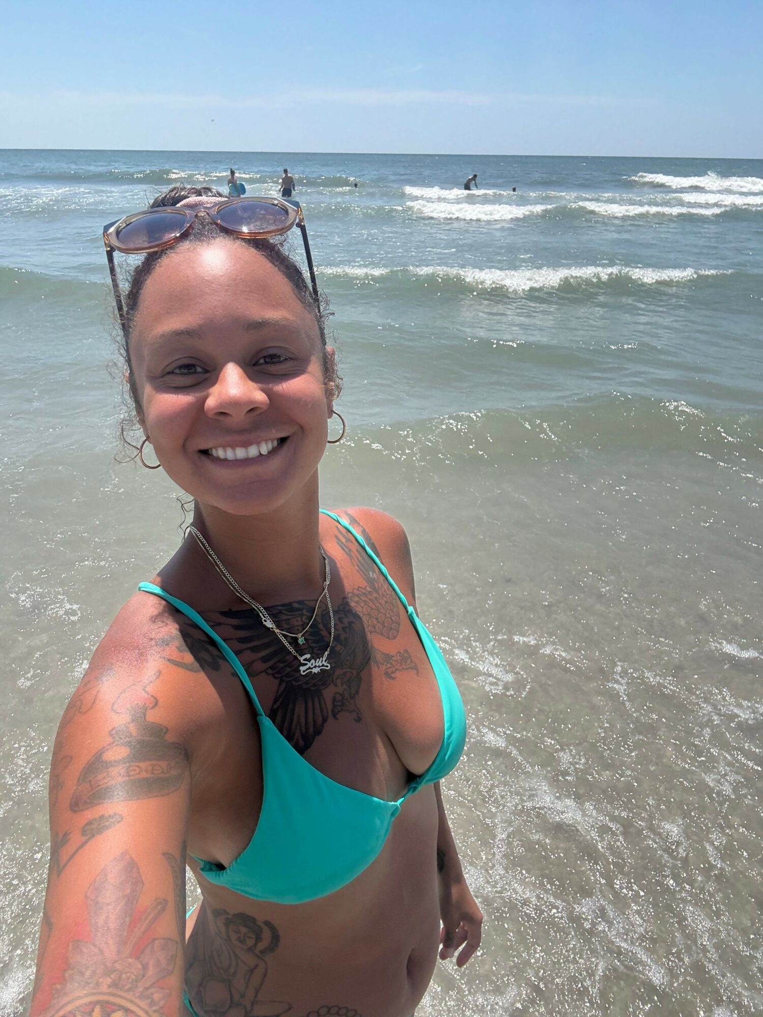 Smiling woman in a turquoise swimsuit at the beach, with ocean waves in the background.