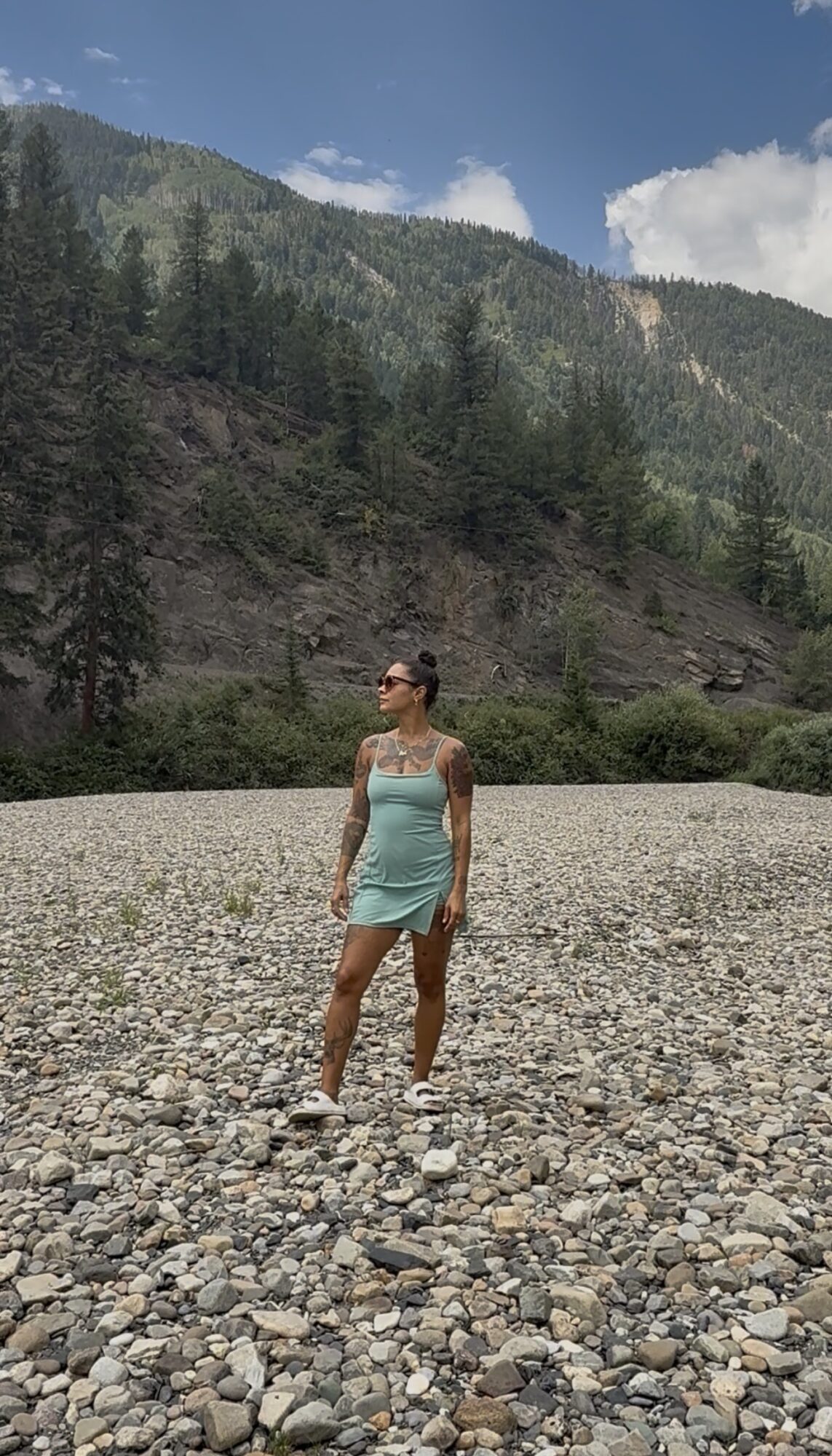 Woman in a light blue dress standing on a rocky terrain with mountains and trees in the background.