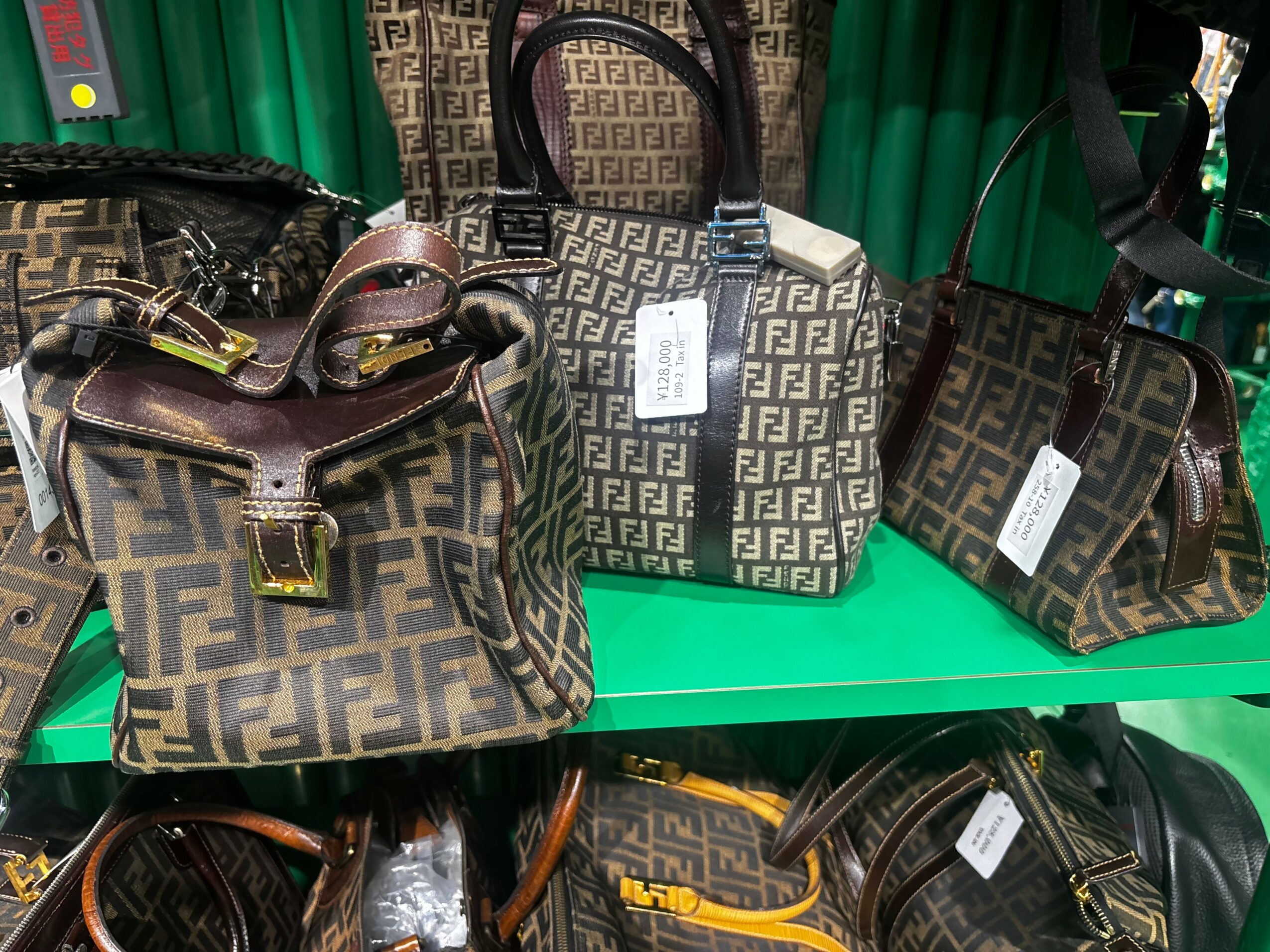 Three designer bags with a repeating pattern, displayed on a green shelf, with tags attached.