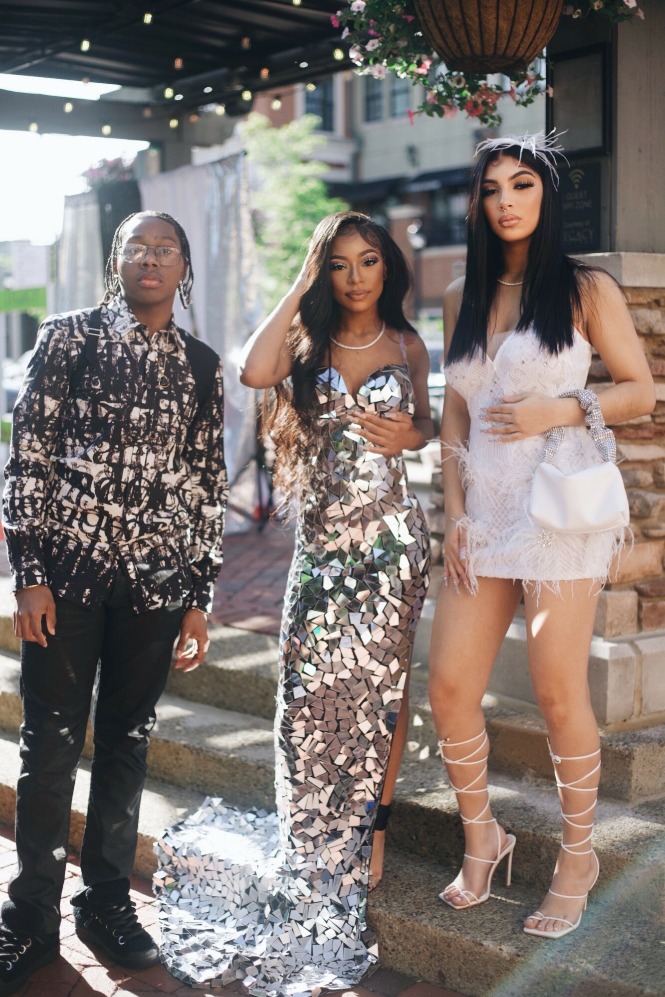Three women standing on a city sidewalk, dressed in shiny, fashionable outfits, with buildings and street in background.