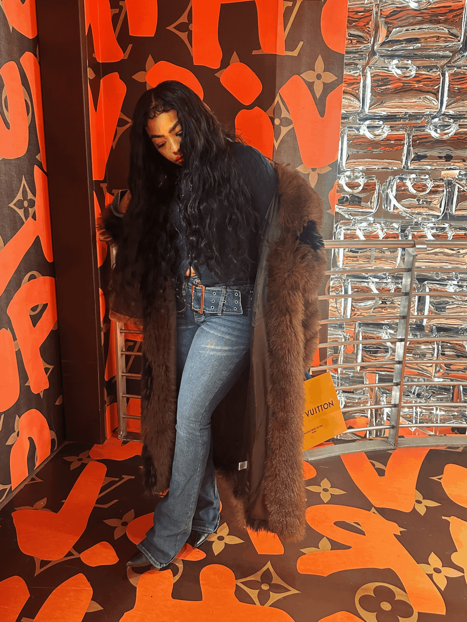 Woman with long dark hair wearing a fur coat and jeans, standing indoors against a patterned wall and reflective surface.