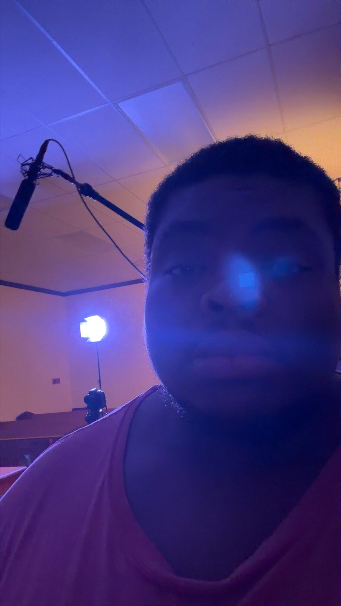 Person with short hair in a dimly lit room with blue and orange lighting, microphone hanging from ceiling, and a bright light in the background.