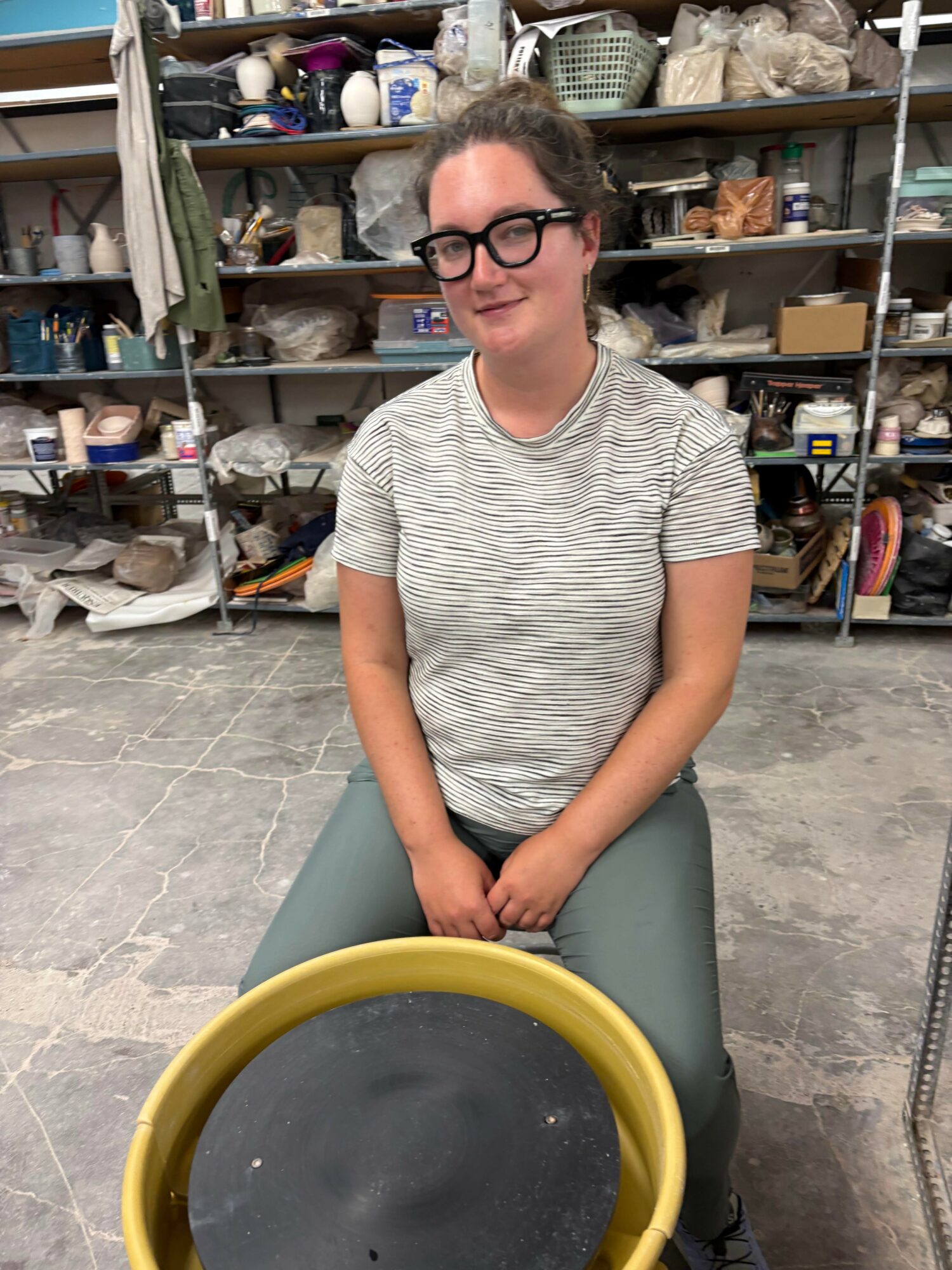 Person sitting on a stool in a storage room with shelves filled with various items behind them.