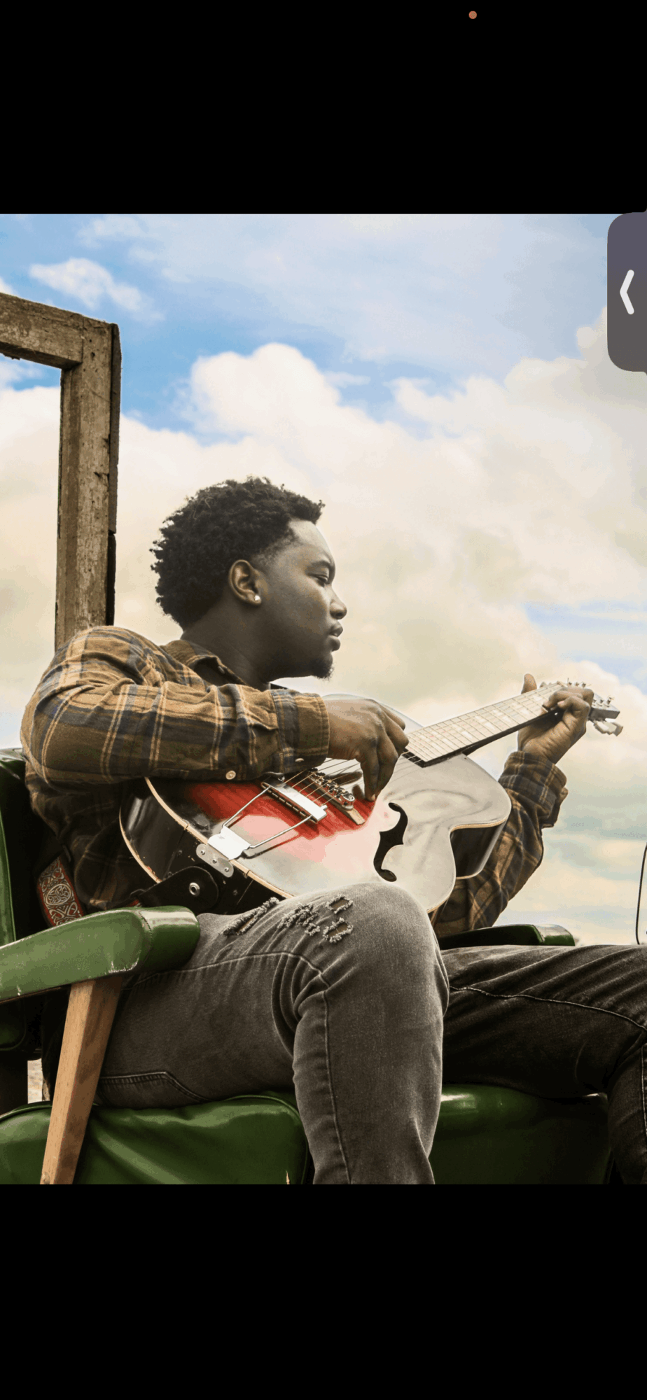 Young man playing guitar outdoors on a green bench, with a cloudy sky background.
