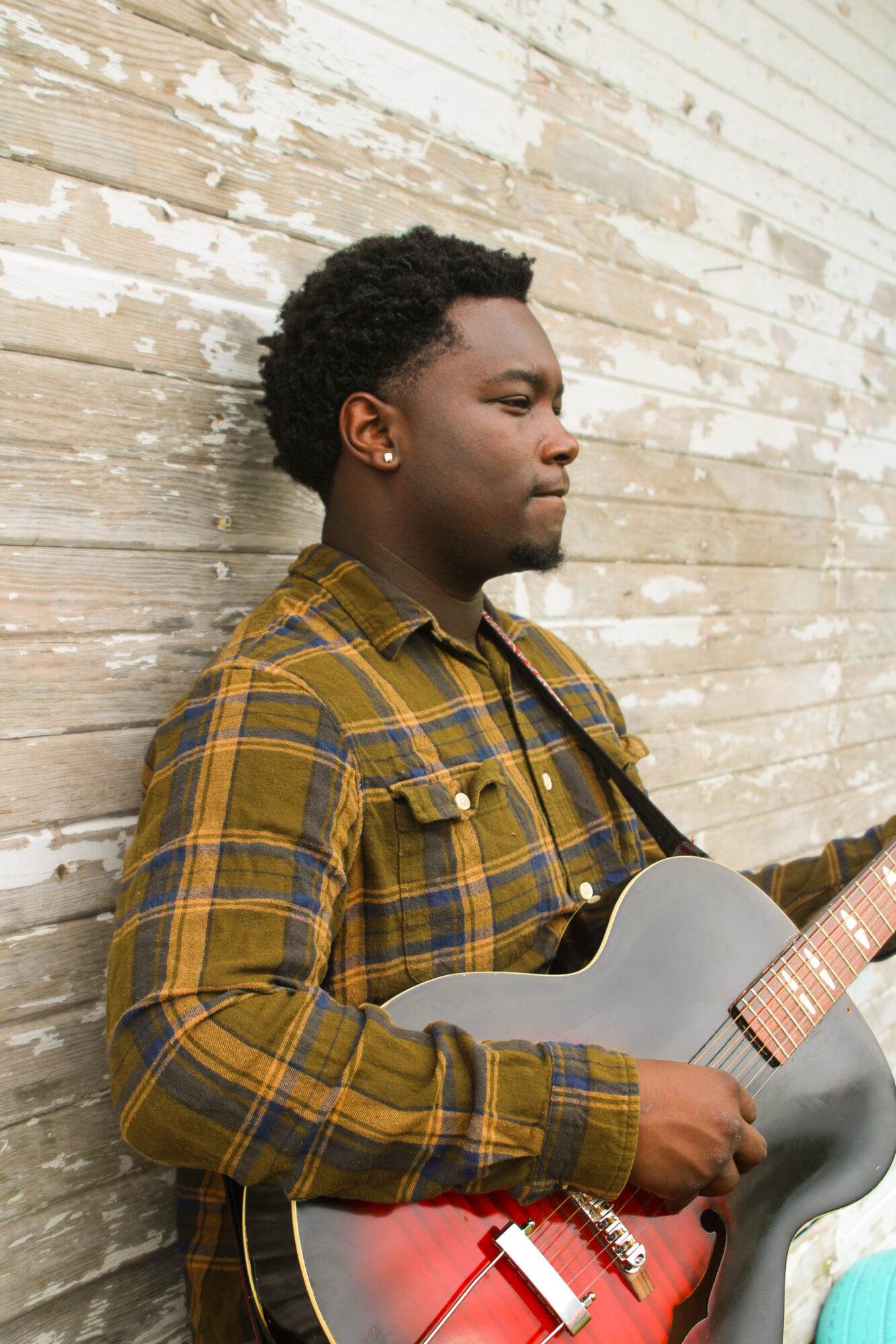 Young man with curly hair in plaid shirt holding a guitar, standing against a weathered wooden wall.
