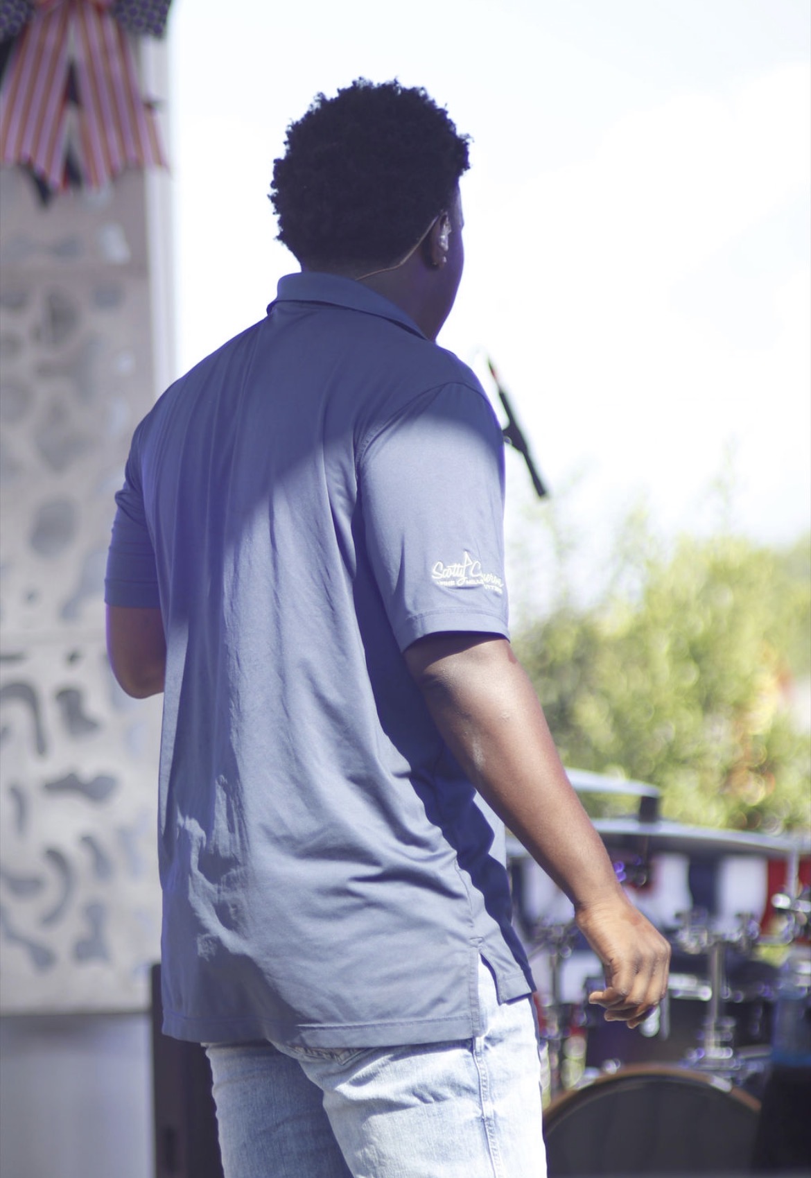Person with curly hair wearing a blue shirt and light pants, standing outdoors near a DJ setup, facing away from the camera.