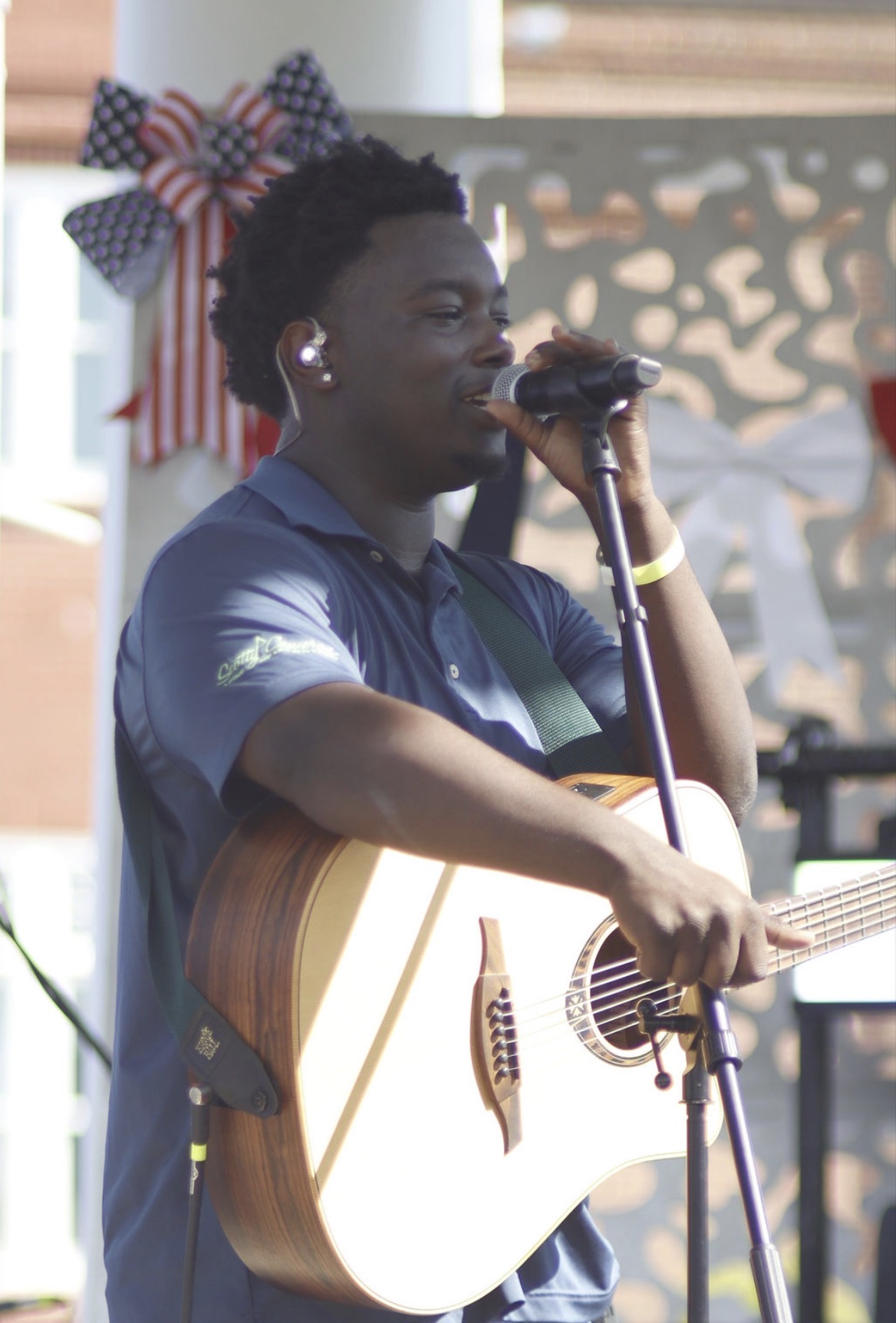 Person singing into microphone while holding an acoustic guitar, outdoors with decorative bow in background.