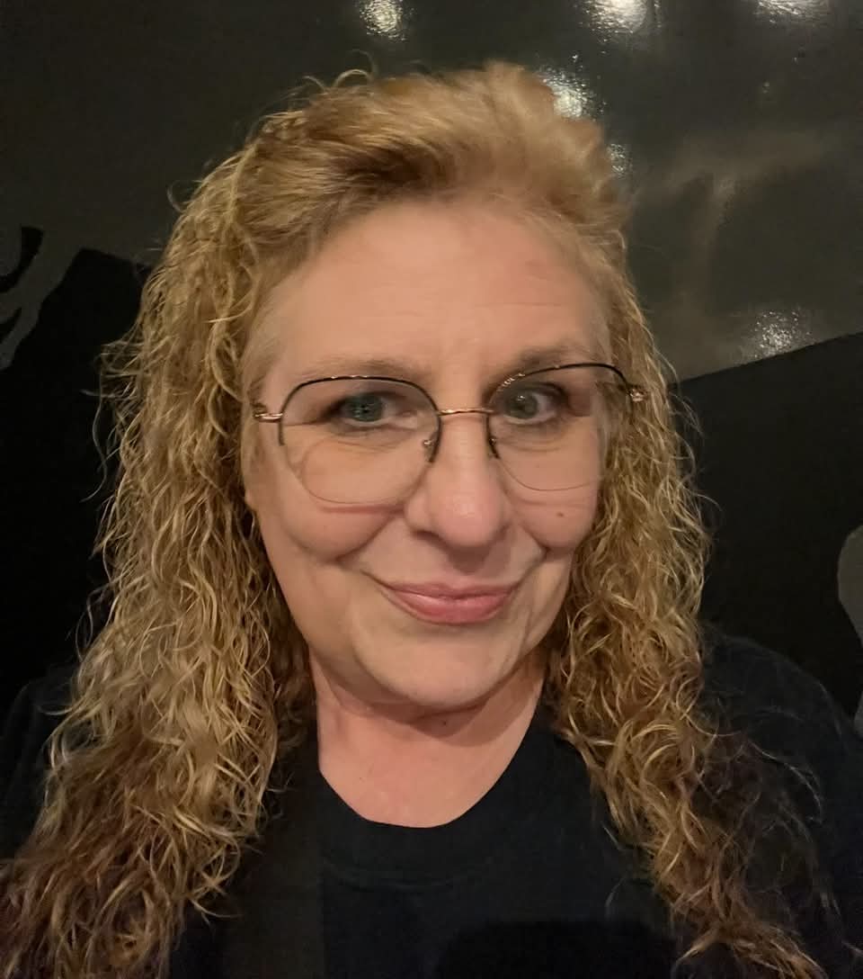 Woman with curly blonde hair and glasses smiling at camera, dark background