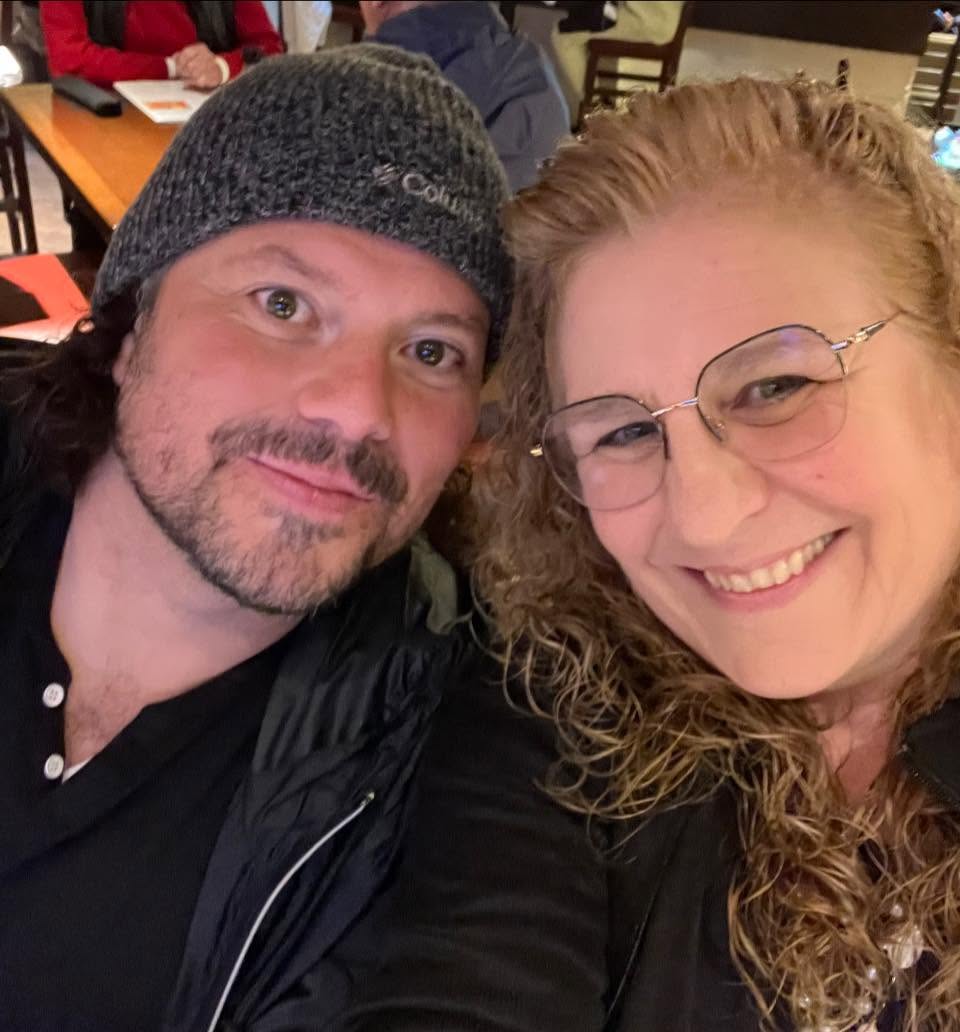 Two people smiling, one wearing a gray beanie and the other with curly hair and glasses, in a restaurant setting.