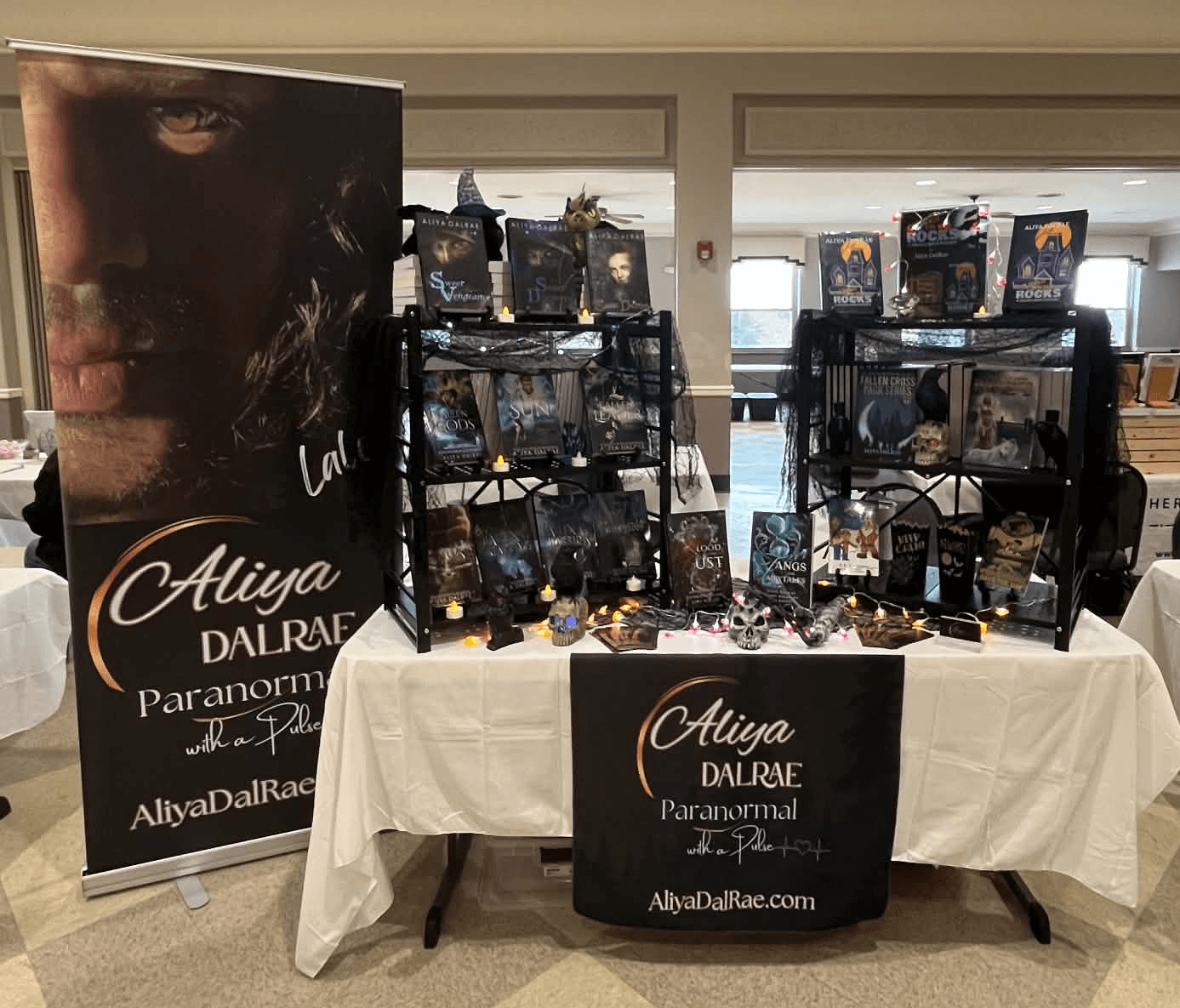 Display table with books, jewelry, and promotional materials, with a large banner featuring a woman's face and text about Aliya Dalrae Paranormal.