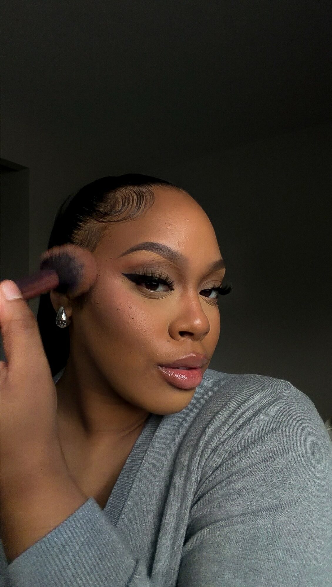 Woman with makeup and earrings, holding a makeup brush near her face, wearing a gray top.