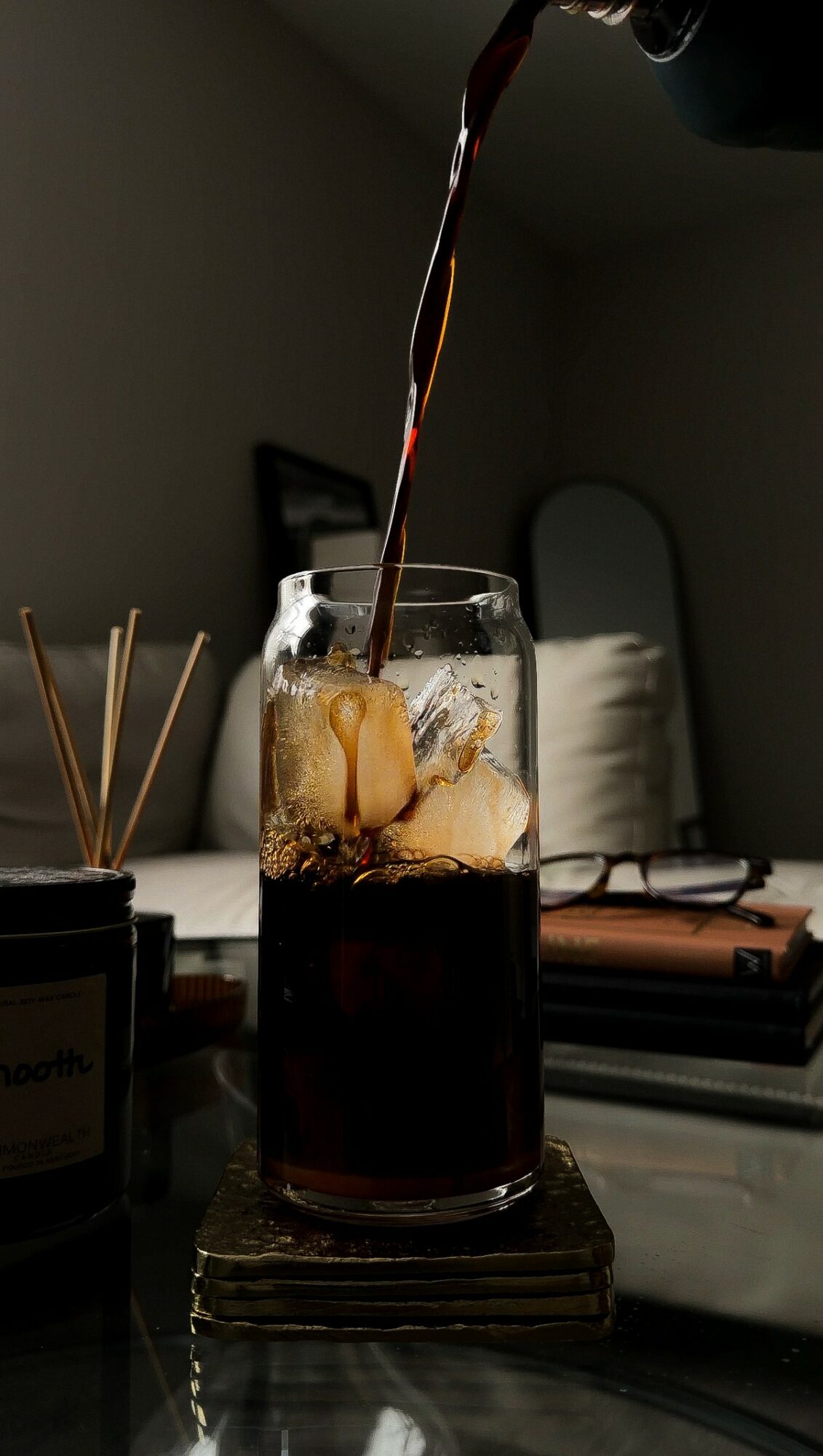 Pouring dark soda into a glass with ice, on a table with various objects in the background.